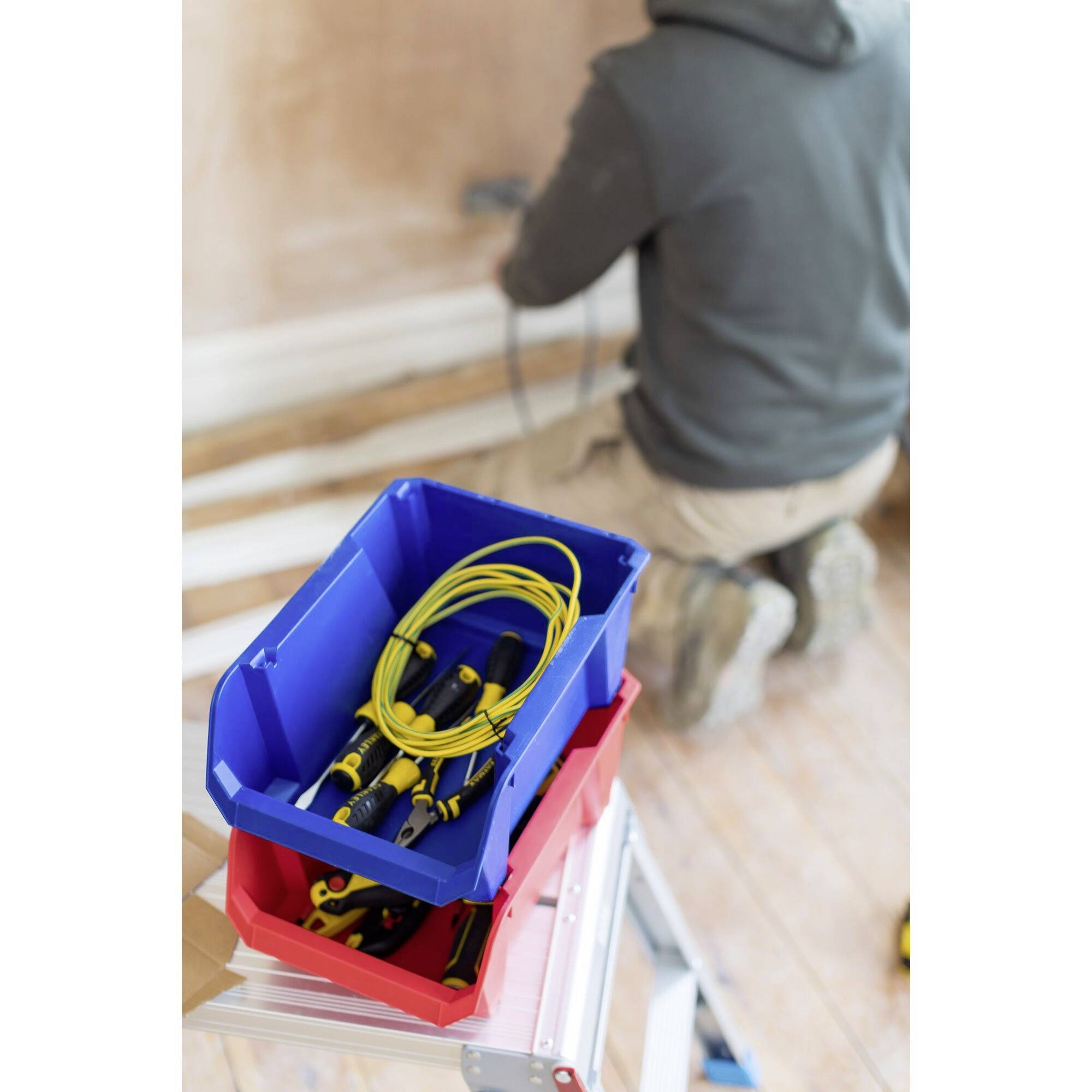 Una persona sta lavorando inginocchiata su una presa a muro. In primo piano, un cassetta degli attrezzi blu con cavi gialli è posizionata su una scala.