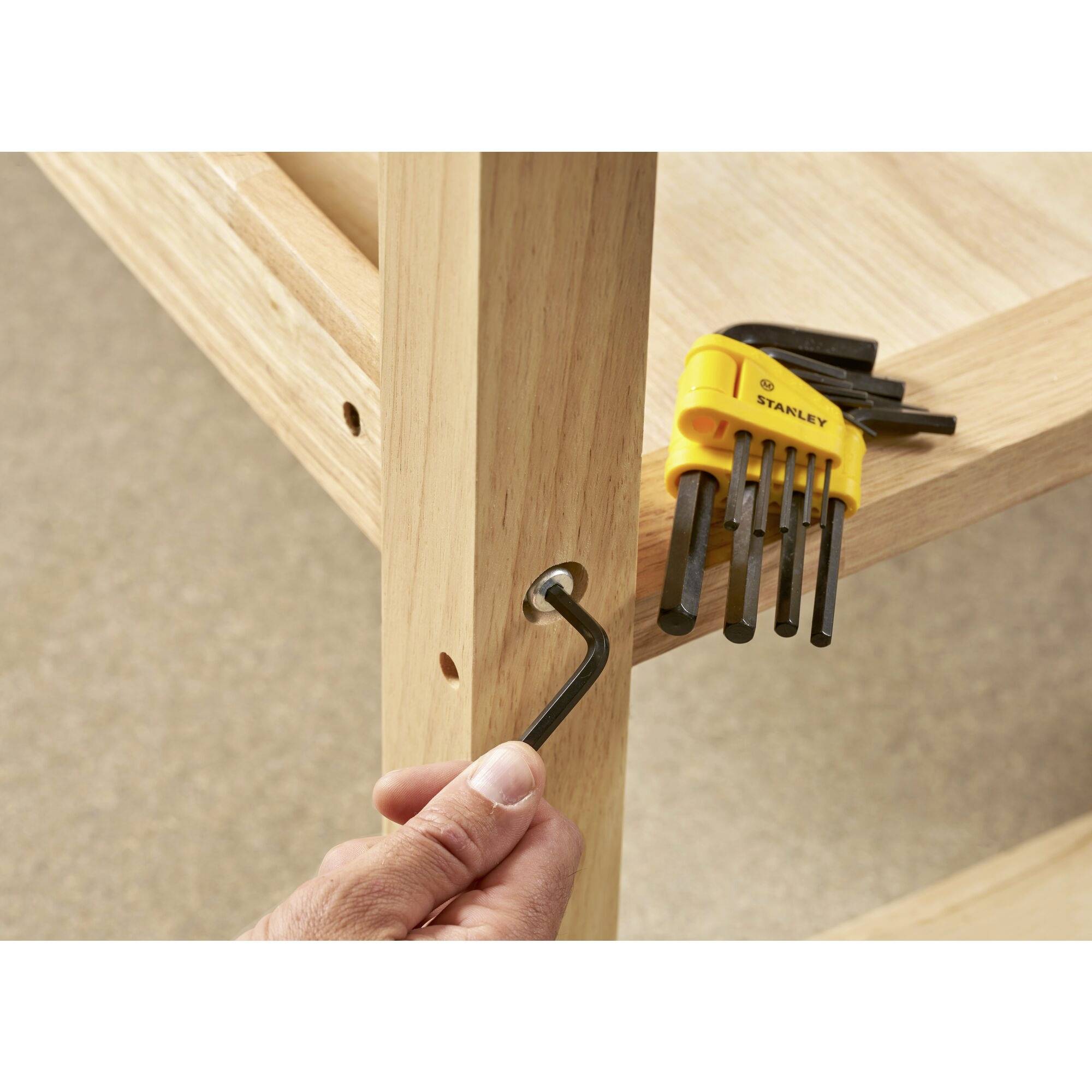 Una mano sta montando un mobile in legno utilizzando una chiave esagonale. Un set di chiavi esagonali in un supporto giallo è posizionato nelle vicinanze.