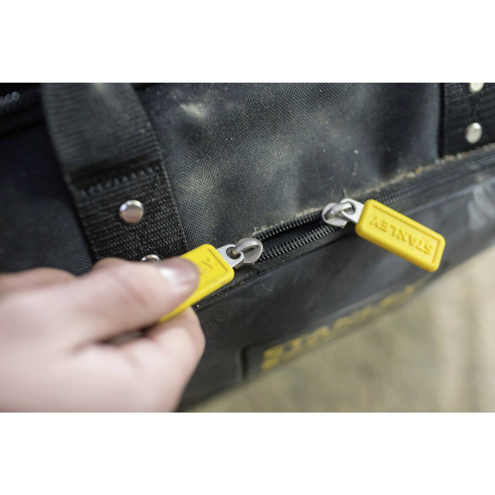 Una mano apre una borsa degli attrezzi nera con tirette gialle su uno sfondo polveroso.