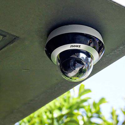 Telecamera di sorveglianza montata sul soffitto, circondata da foglie verdi sullo sfondo, simbolo di sicurezza e controllo.