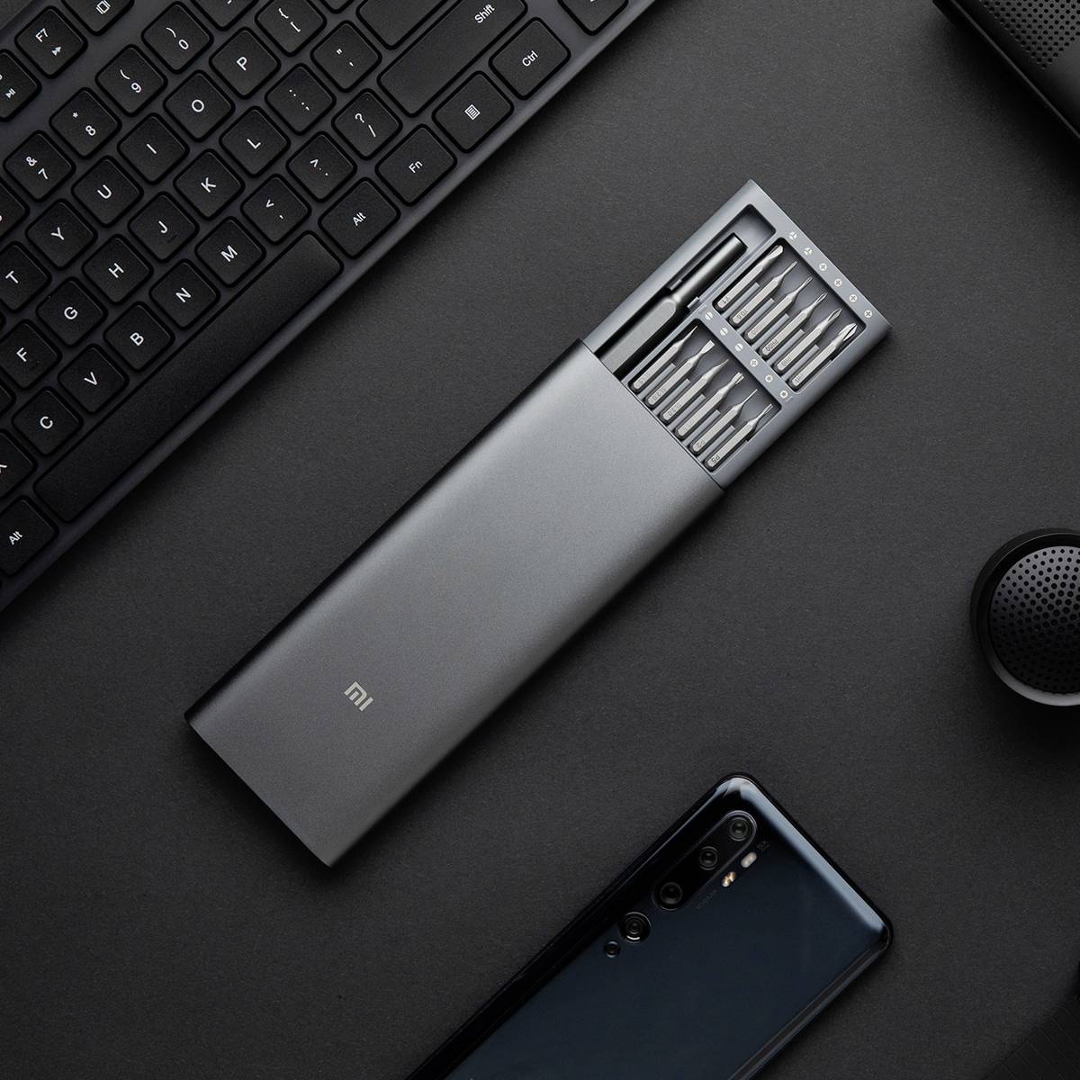 Un set di cacciaviti con vari bit è posizionato accanto a una tastiera e uno smartphone su uno sfondo scuro.