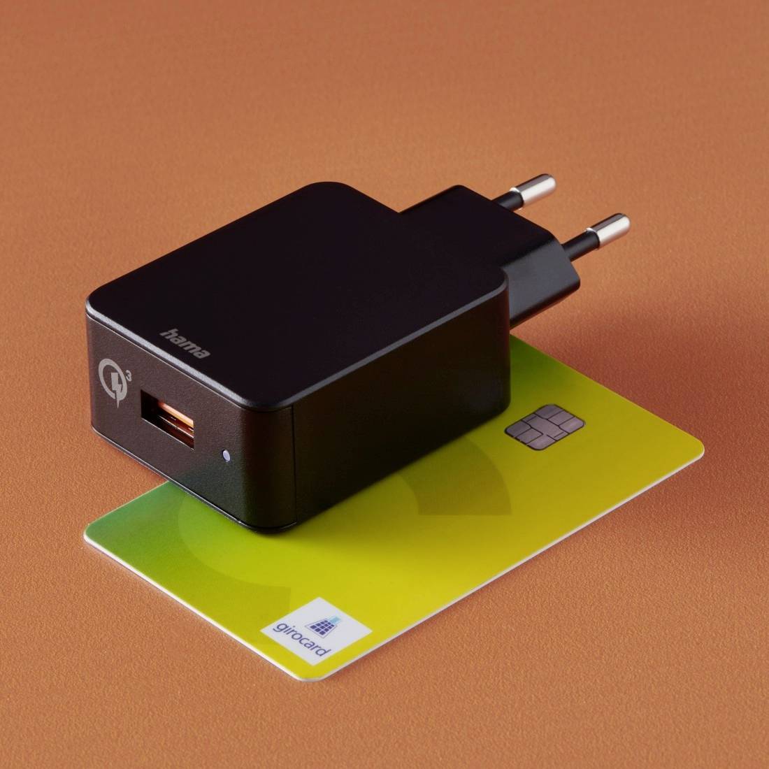 Un caricabatterie USB nero è poggiato su una carta Girocard gialla con chip, su uno sfondo arancione.