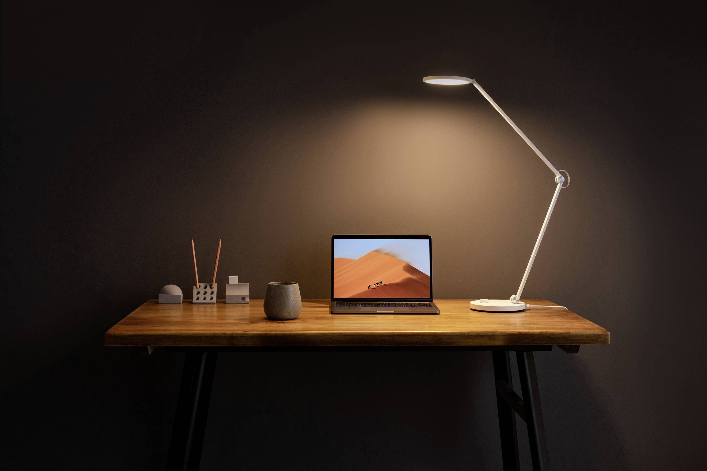Un tavolo minimale con un laptop aperto, una lampada da tavolo moderna e piccoli accessori da ufficio su una superficie in legno.
