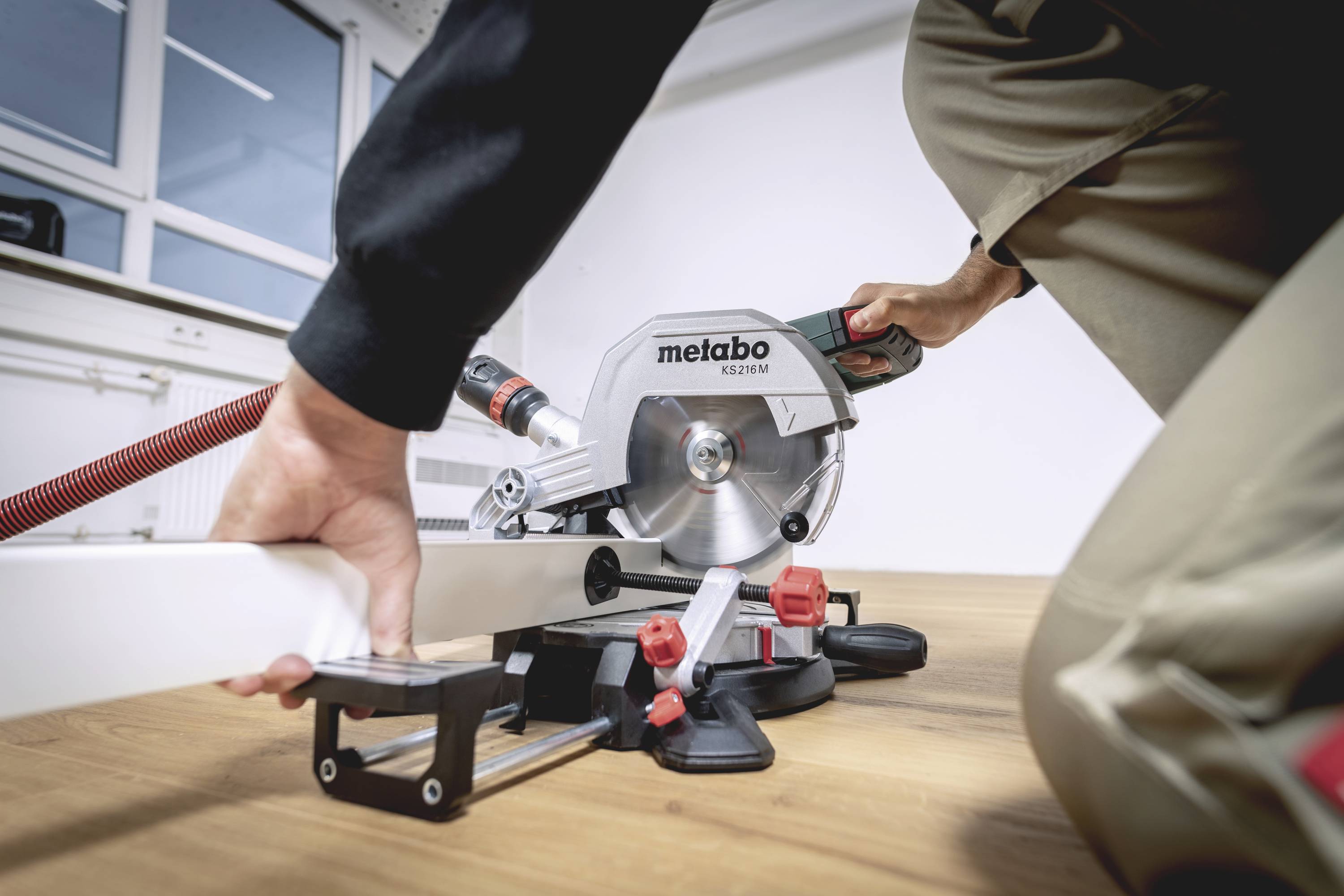 Una persona sta tagliando un pezzo di legno con una sega a troncatore Metabo all'interno di un ambiente. La sega è collegata a un sistema di aspirazione della polvere.