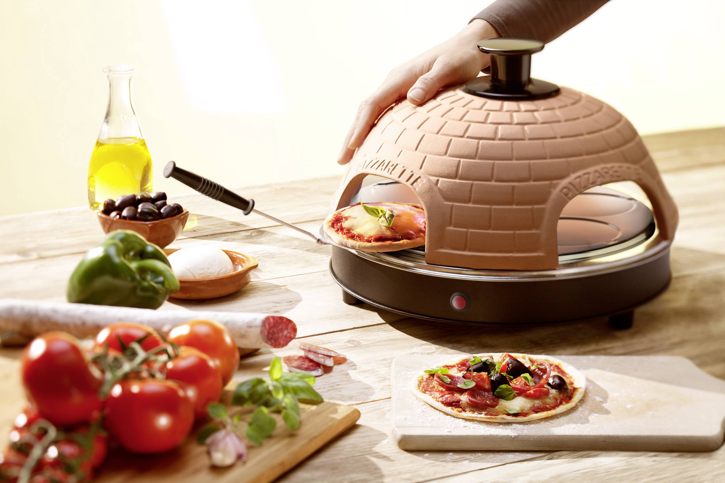 Un mini-forno per pizza con cupola in terracotta, in cui viene cotta una pizza. Ingredienti freschi come pomodori, olive e peperoni sono posizionati accanto.