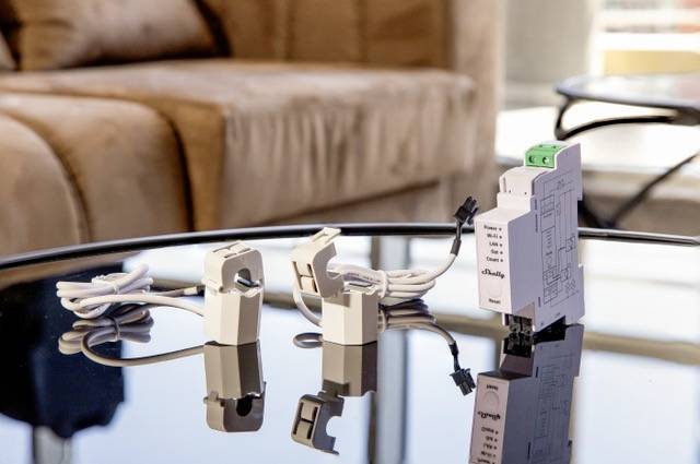 Su un tavolo di vetro sono posizionati due dispositivi elettronici bianchi con cavi, riflessi sulla superficie del tavolo. Sullo sfondo un divano.