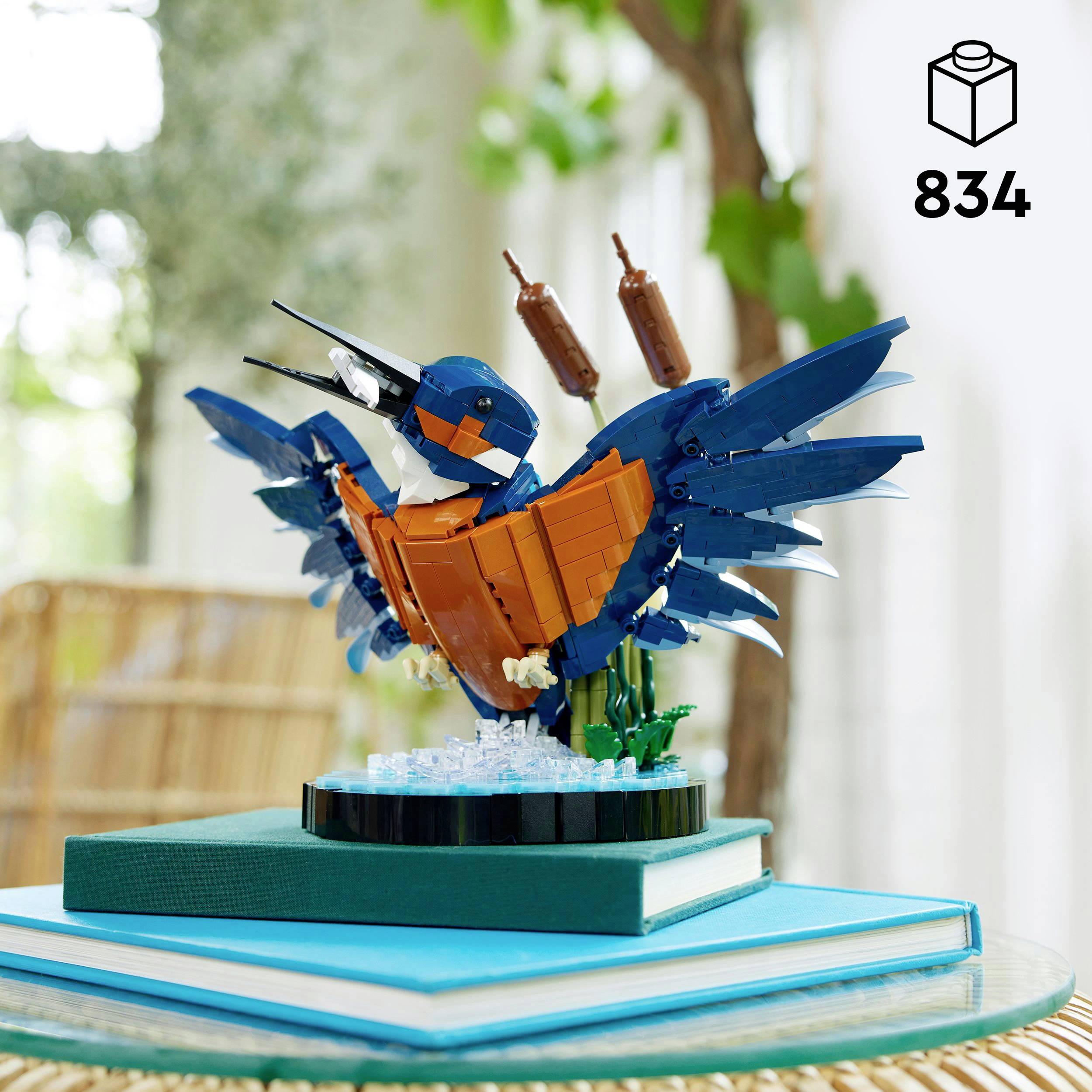 Un colorato uccello Lego è in piedi con le ali spiegate su un piedistallo. Accanto sono raffigurate piante e acqua.