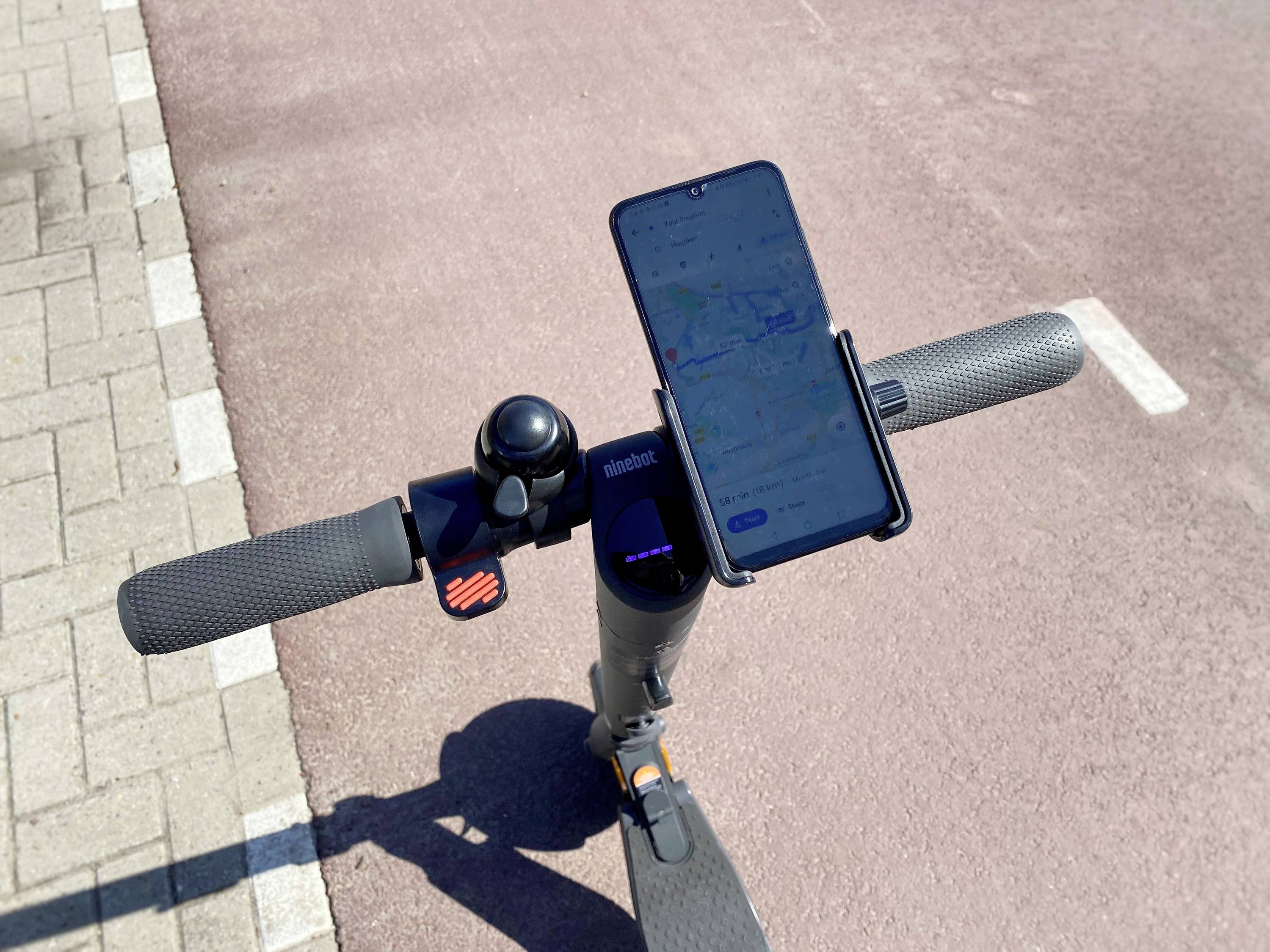 Manubrio di un monopattino elettrico con smartphone fissato che mostra un'app di navigazione. Superficie di pavimentazione e asfalto sullo sfondo.
