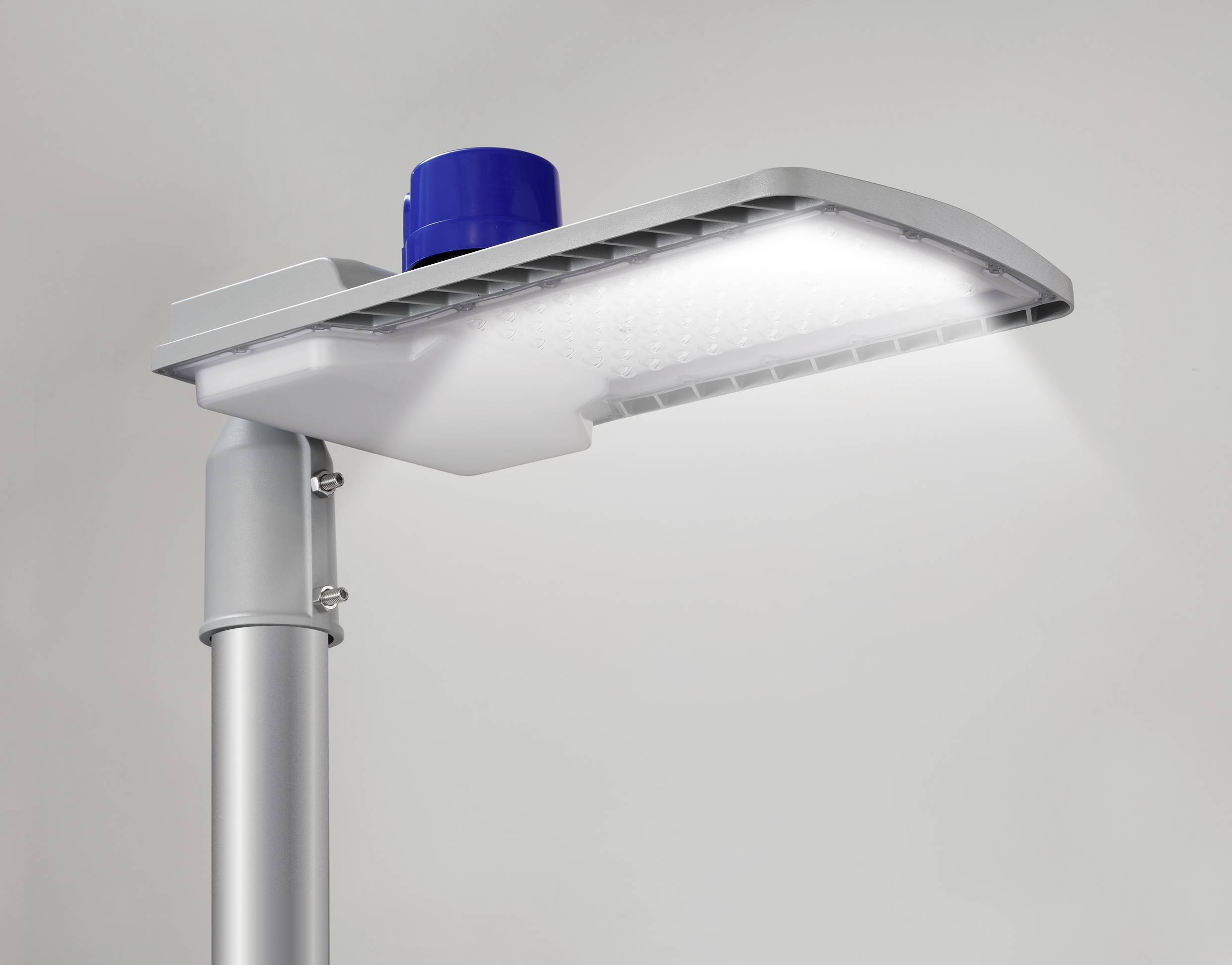 Una moderna lampada stradale a LED illumina intensamente. È fissata a un palo metallico grigio, con un coperchio blu in cima.