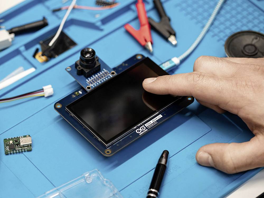 Una mano tocca lo schermo di un microcontrollore su un tavolo blu, circondato da componenti elettronici e strumenti.