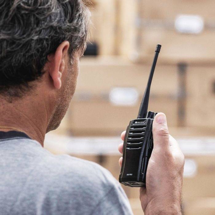 Una persona tiene un walkie-talkie mentre parla rivolto verso scatole impilate. La scena suggerisce attività logistiche o di magazzino.