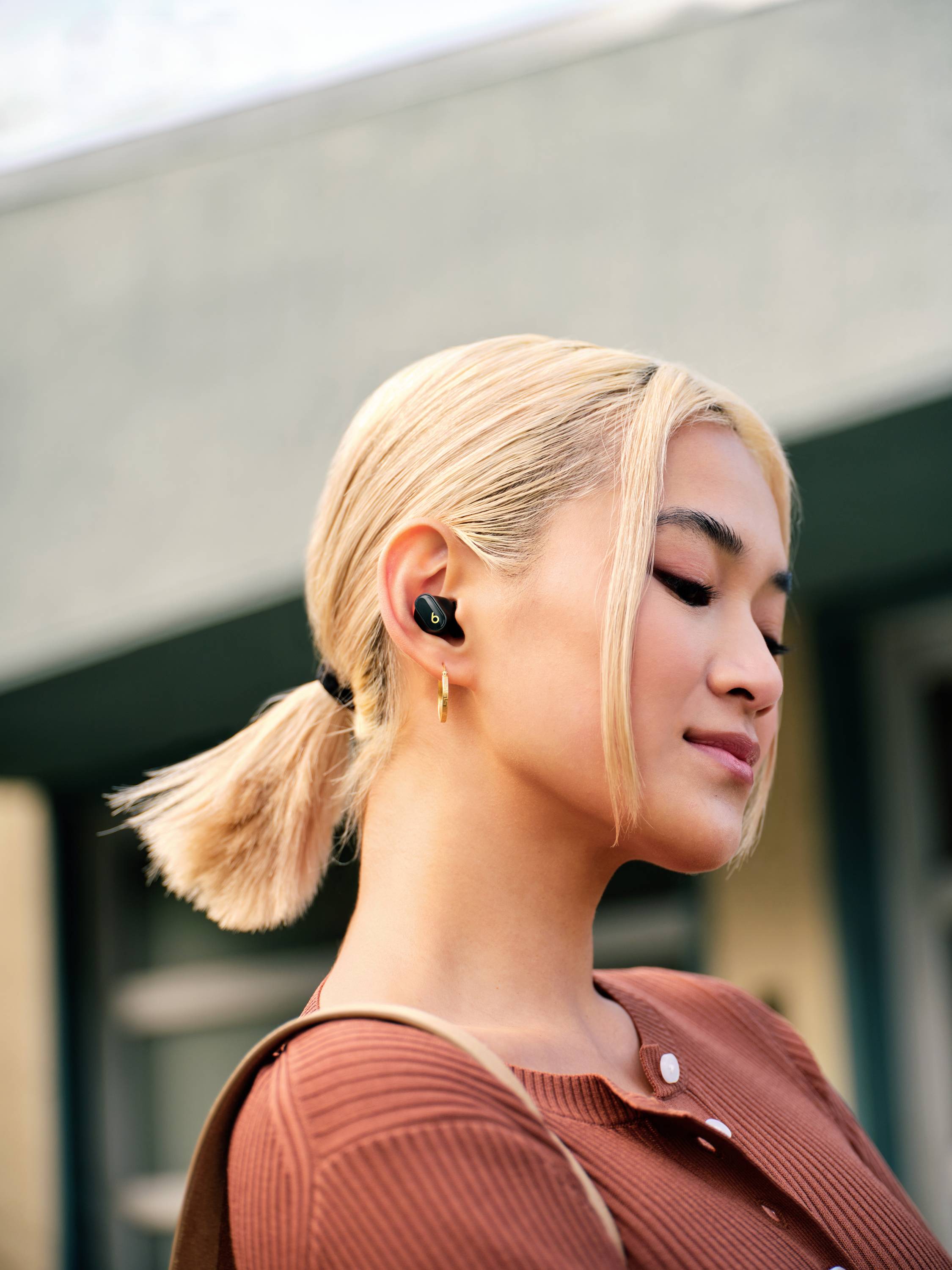 Una persona con capelli biondi indossa auricolari wireless ed è in piedi davanti a un edificio sfocato. Indossa una maglia marrone.