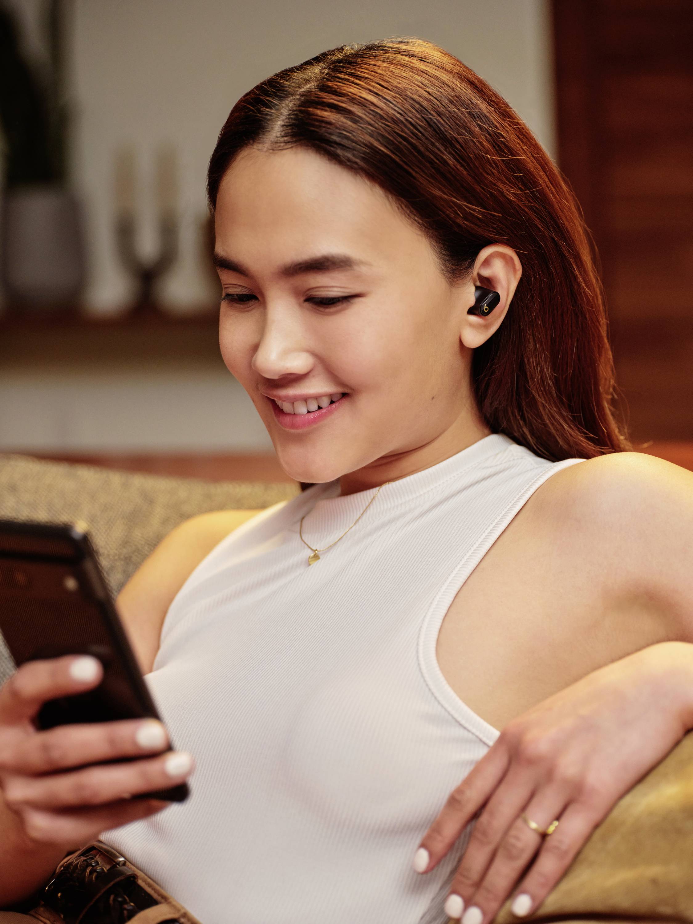 Donna seduta sul divano, sorride mentre guarda il suo smartphone. Indossa cuffie wireless. Soggiorno sullo sfondo.