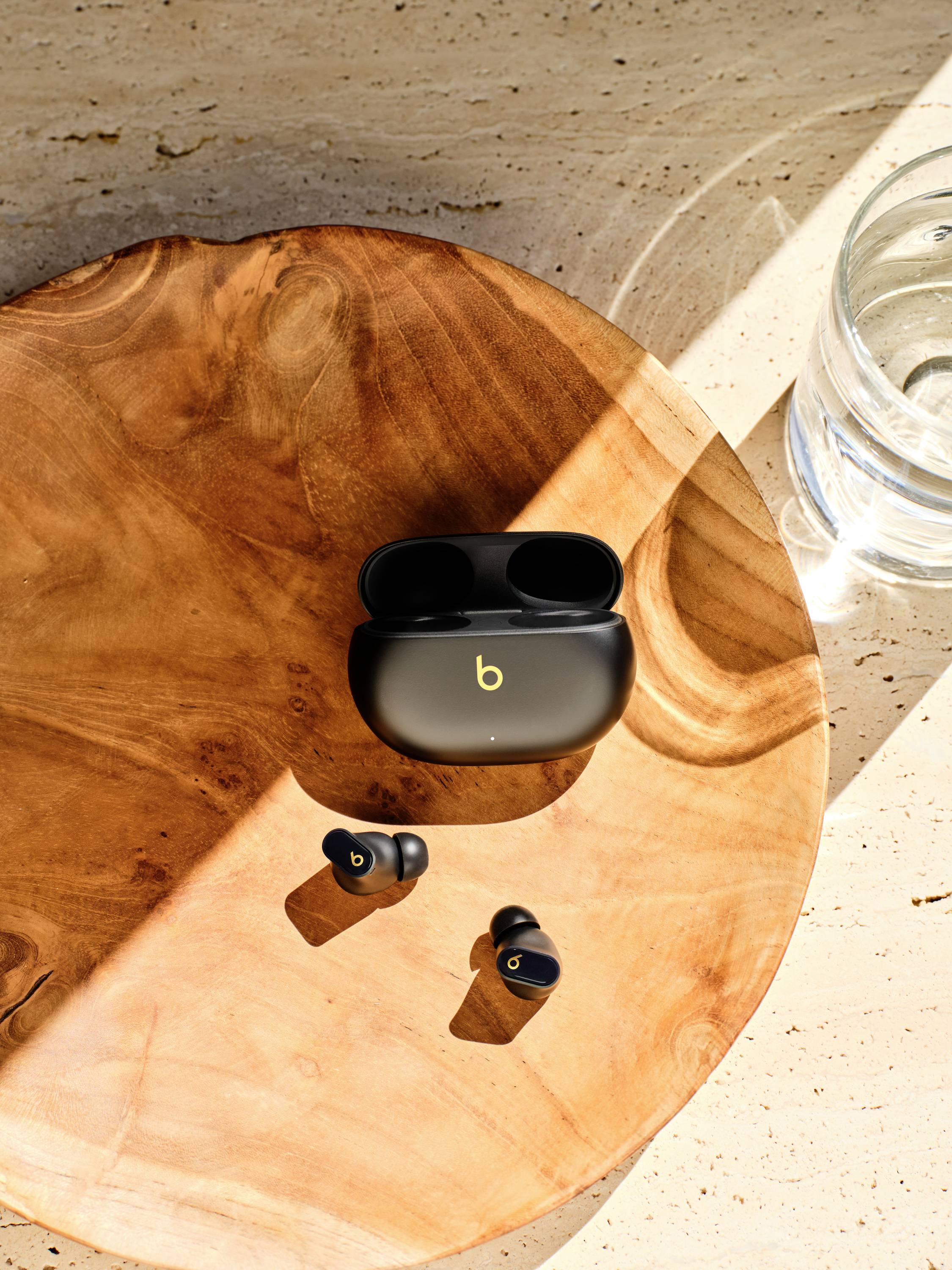 Un paio di auricolari wireless e il relativo astuccio di ricarica aperto sono posizionati su un tavolo rotondo in legno, accanto a un bicchiere d'acqua, sotto la luce del sole.