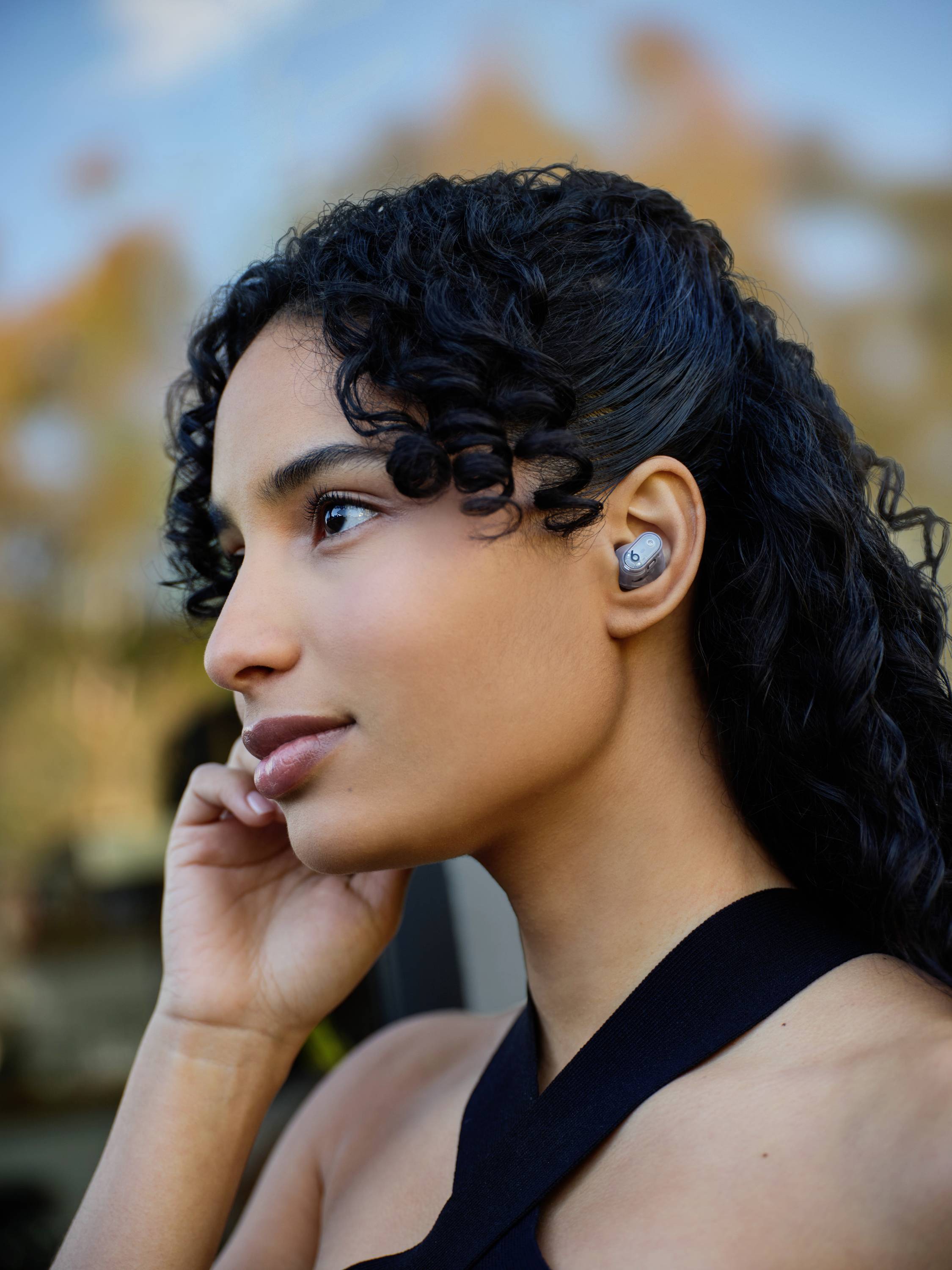 Una persona con capelli ricci indossa auricolari wireless e guarda di lato, sullo sfondo una natura sfocata.