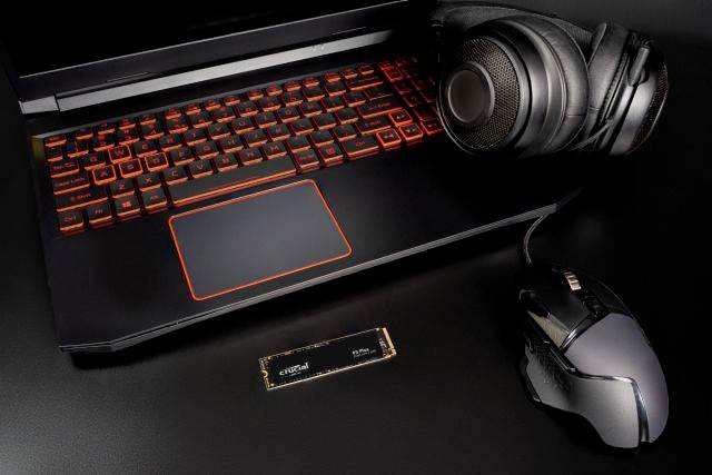 Laptop con illuminazione tastiera rossa, cuffie, mouse per computer e SSD M.2 NVMe su tavolo nero. Accessori tecnici per gaming.