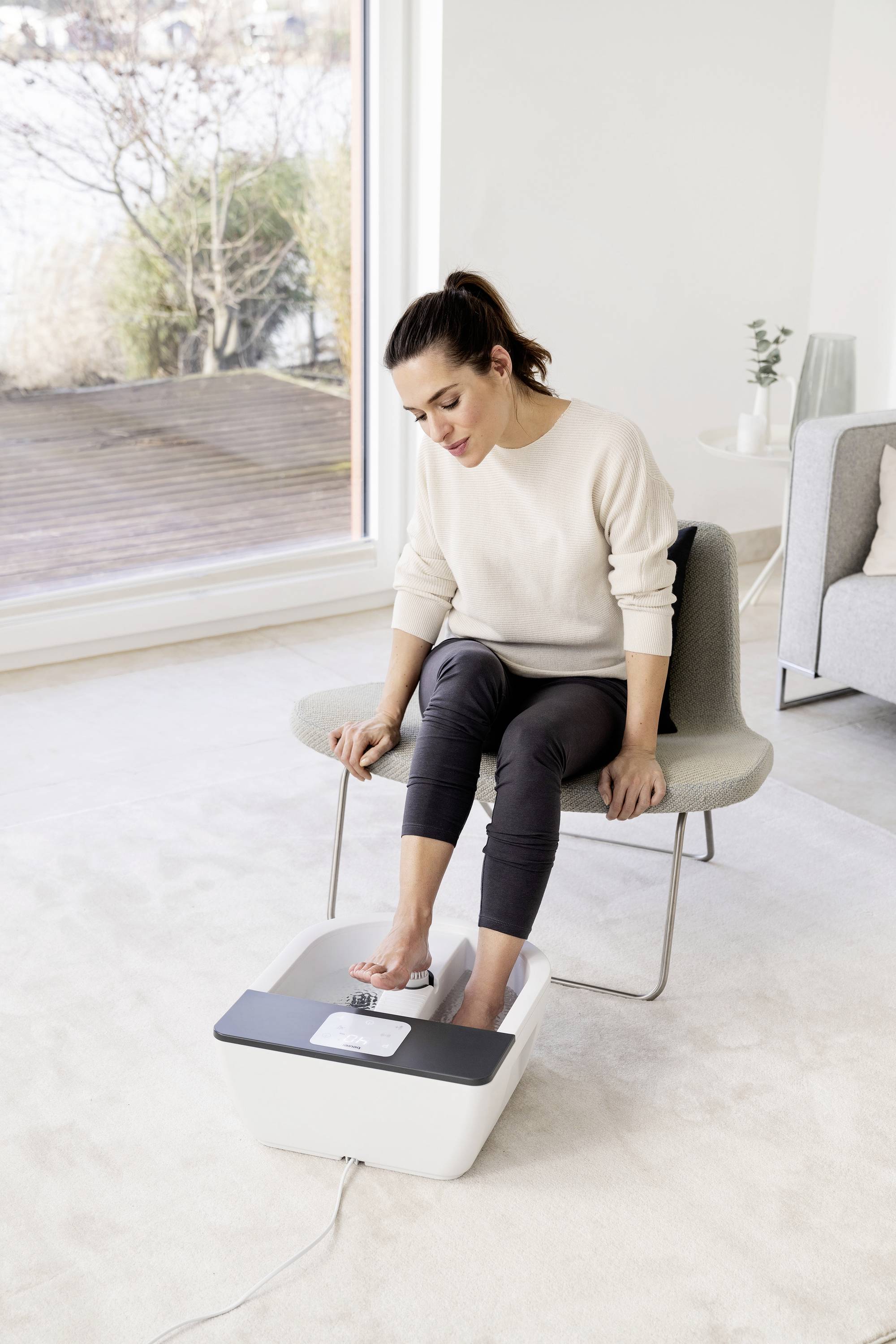 Una donna è seduta in un soggiorno moderno e utilizza un dispositivo per il bagno dei piedi. Indossa abiti casual e si sta rilassando con i piedi.
