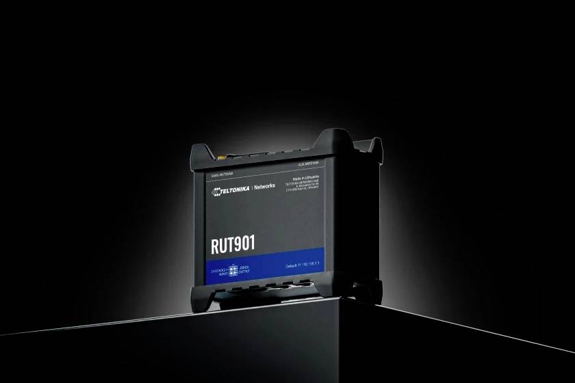 Un dispositivo di telecomunicazione nero con la scritta 'RUT901' è posizionato su un piedistallo. Sullo sfondo scuro, il dispositivo risalta in primo piano.
