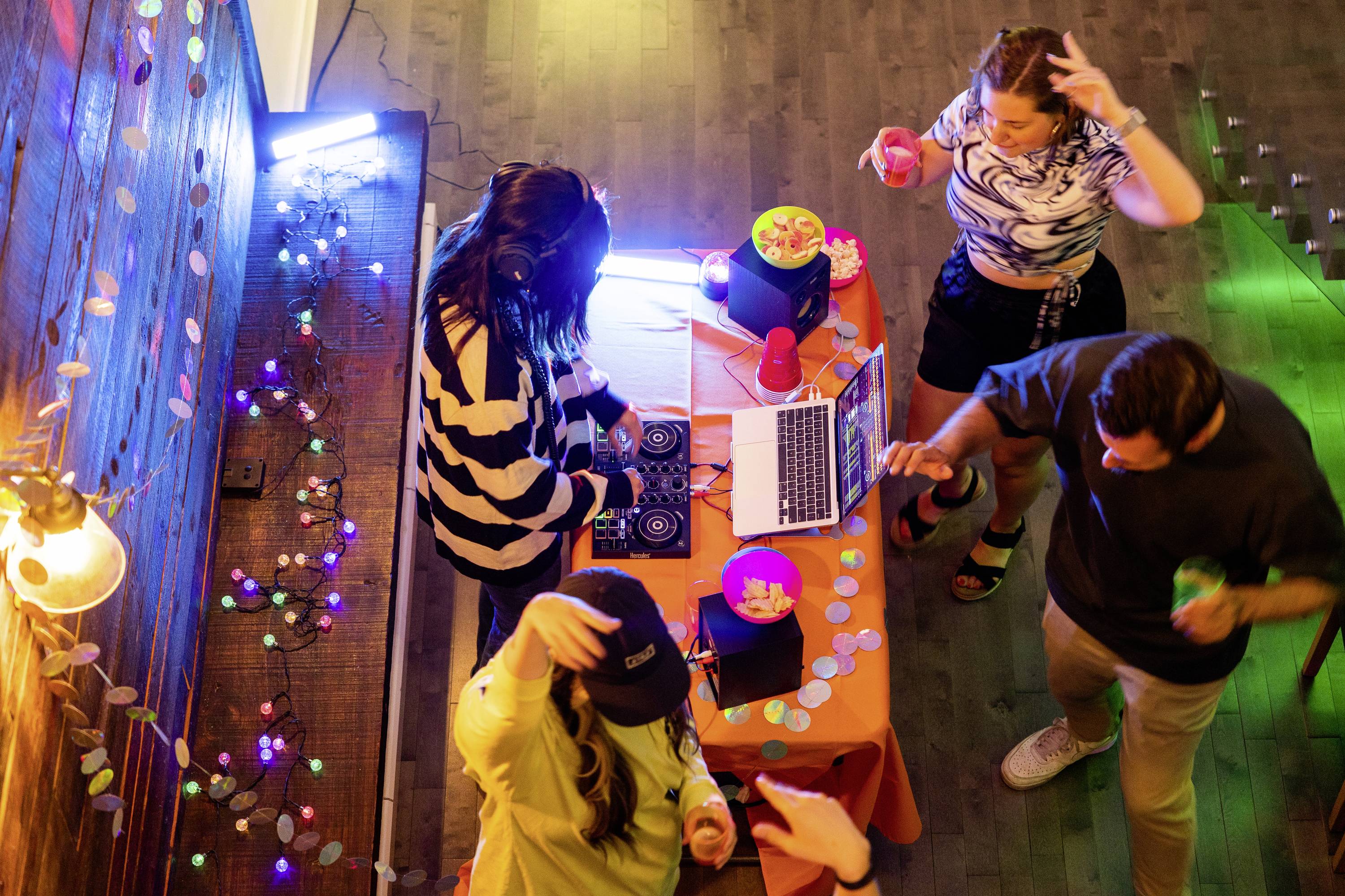 Un gruppo di giovani balla intorno a un tavolo con un controller DJ e un laptop. Luci colorate creano un'atmosfera vivace da festa.