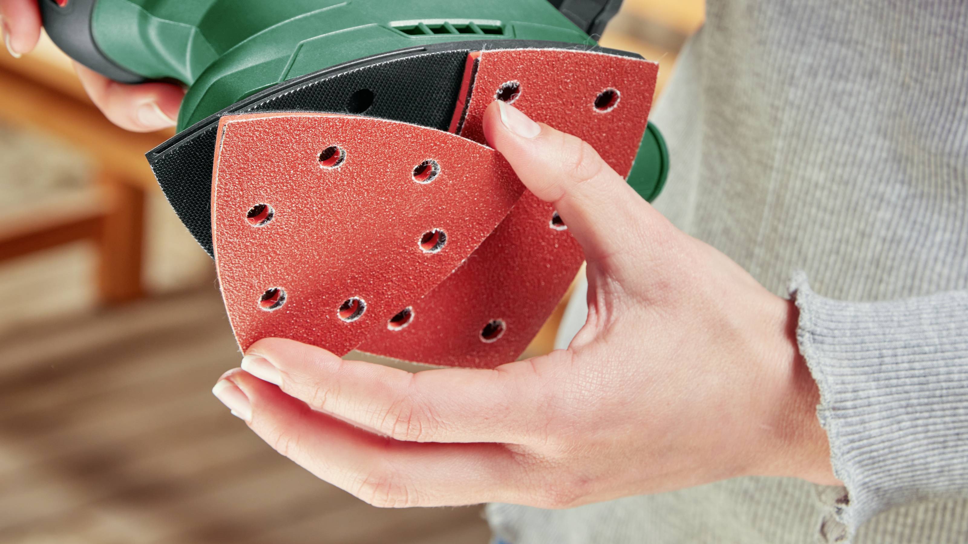 Una persona tiene un utensile elettrico verde con un pad rosso per levigare, pronta a iniziare la levigatura. Il pad presenta fori per l'aspirazione della polvere.