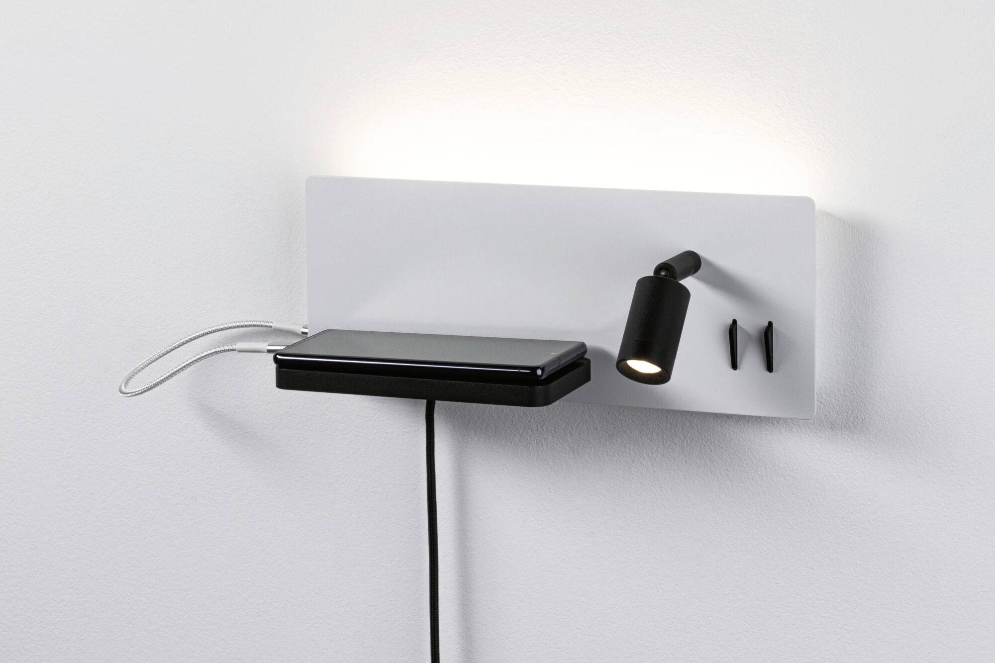 Lampada da parete con scaffale integrato che sorregge uno smartphone; sotto di esso è presente un caricabatterie. Un piccolo faretto regolabile è fissato accanto.
