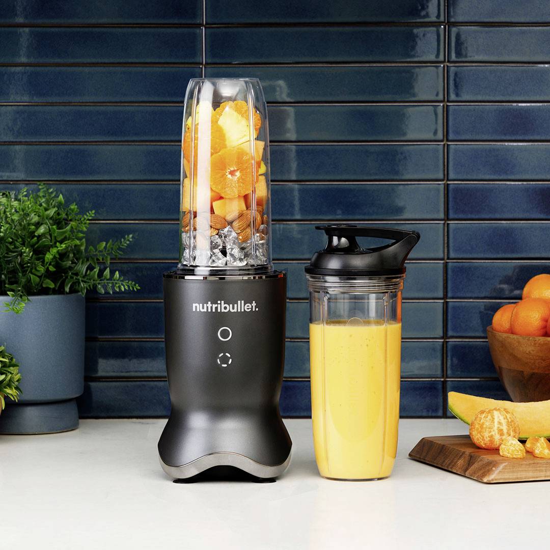 Un frullatore è posizionato su un piano di lavoro della cucina, riempito con pezzi di frutta. Accanto a esso, un bicchiere con uno smoothie fresco, con piante sullo sfondo.
