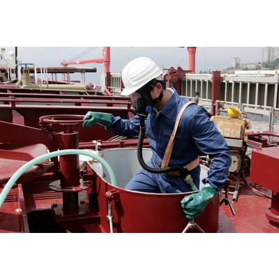 Una persona in abbigliamento protettivo e maschera respiratoria sale in un serbatoio su una nave. Attrezzature di sicurezza e ambiente industriale sono visibili.