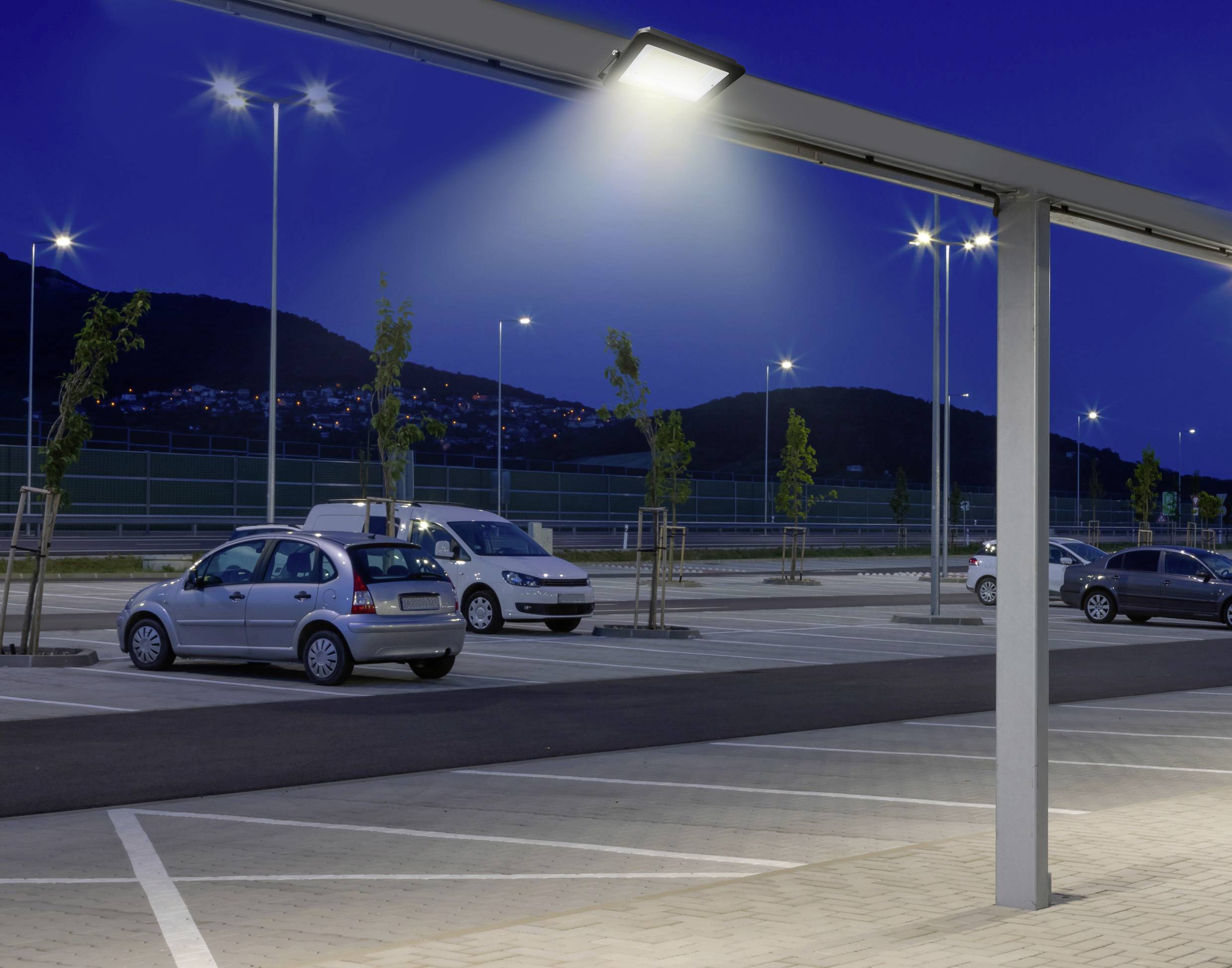 Parcheggio illuminato di notte con diverse auto parcheggiate e, sullo sfondo, un paesaggio collinare con luci.