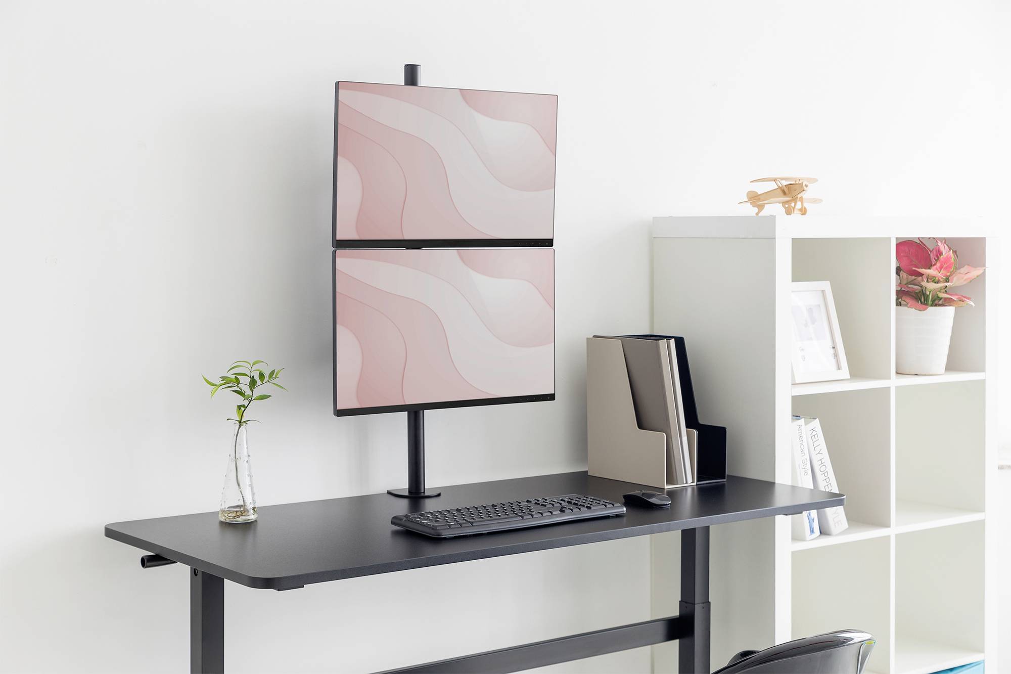 Un moderno scrivania da lavoro in piedi di colore nero con due monitor posizionati uno sopra l'altro. A sinistra una piccola pianta, a destra uno scaffale.