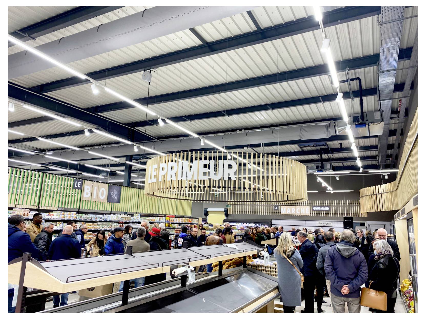 Interno affollato di un supermercato, con clienti raccolti lungo le corsie e intorno alle casse, sotto luci intense al soffitto e grandi cartelli appesi.