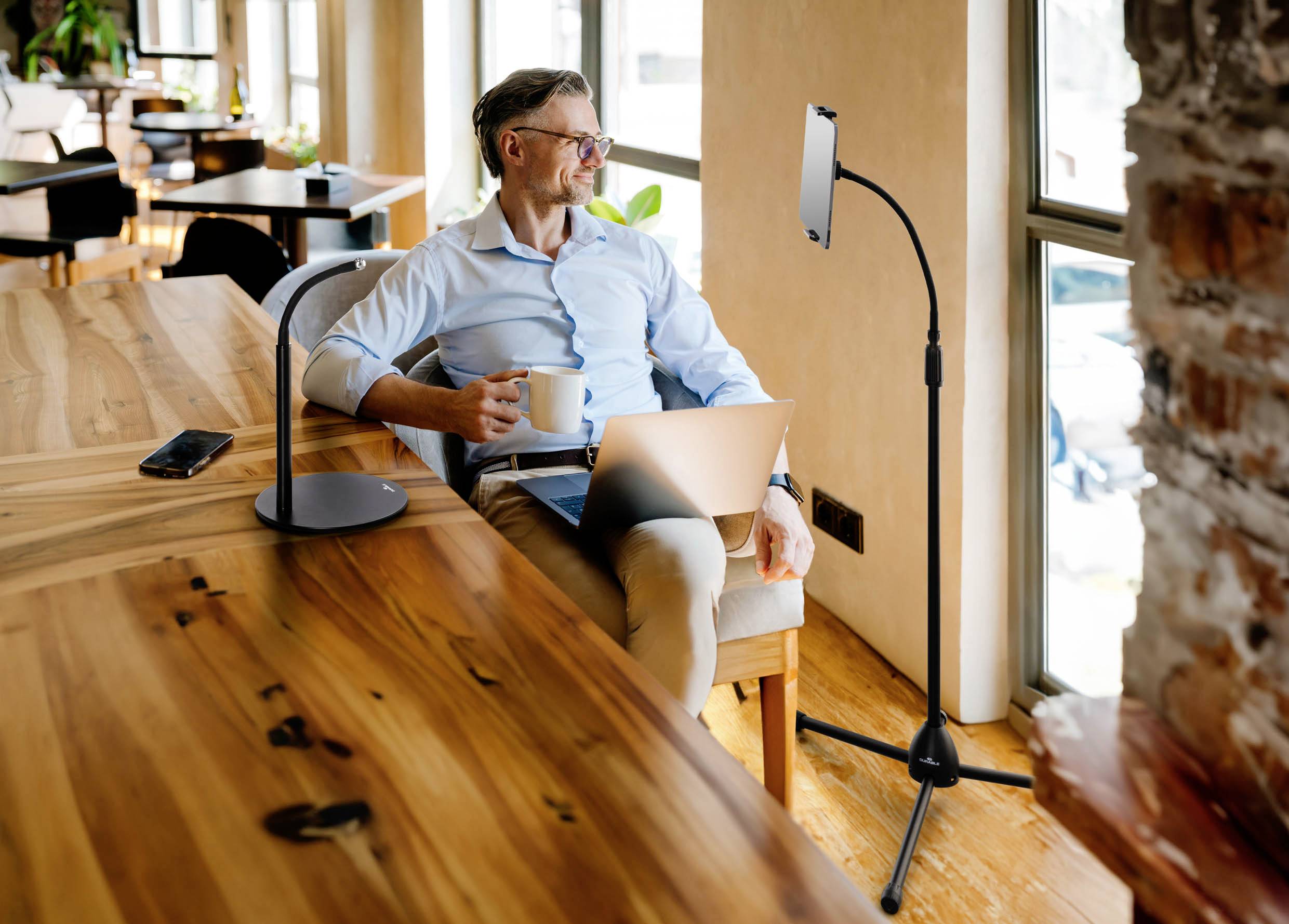 Un uomo è seduto in un caffè, tiene una tazza di caffè e guarda un tablet posizionato su un supporto. Ha un laptop sulle ginocchia.