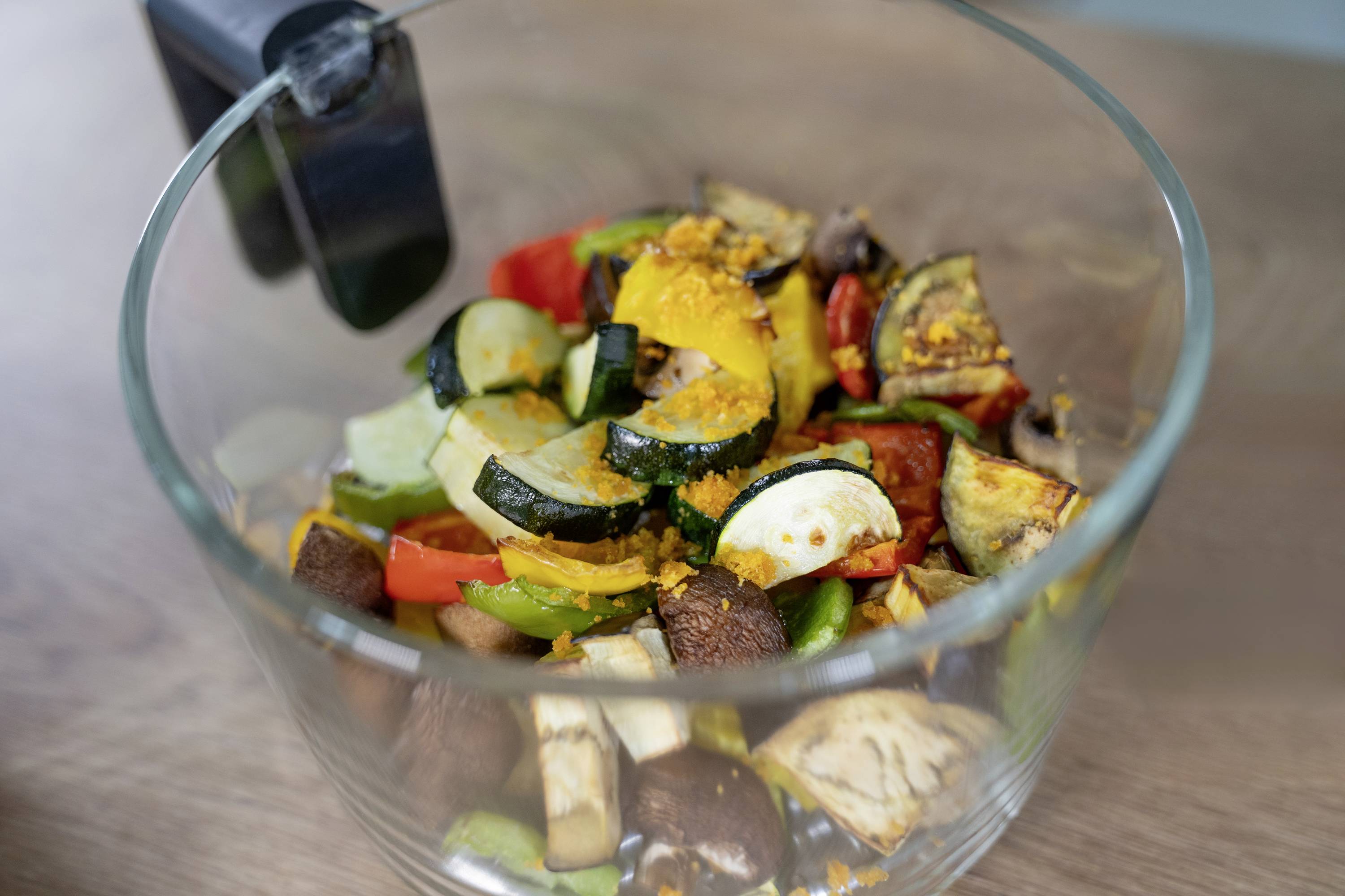 Una ciotola di vetro con verdure fresche tagliate come zucchine, peperoni e funghi champignon su un tavolo di legno.
