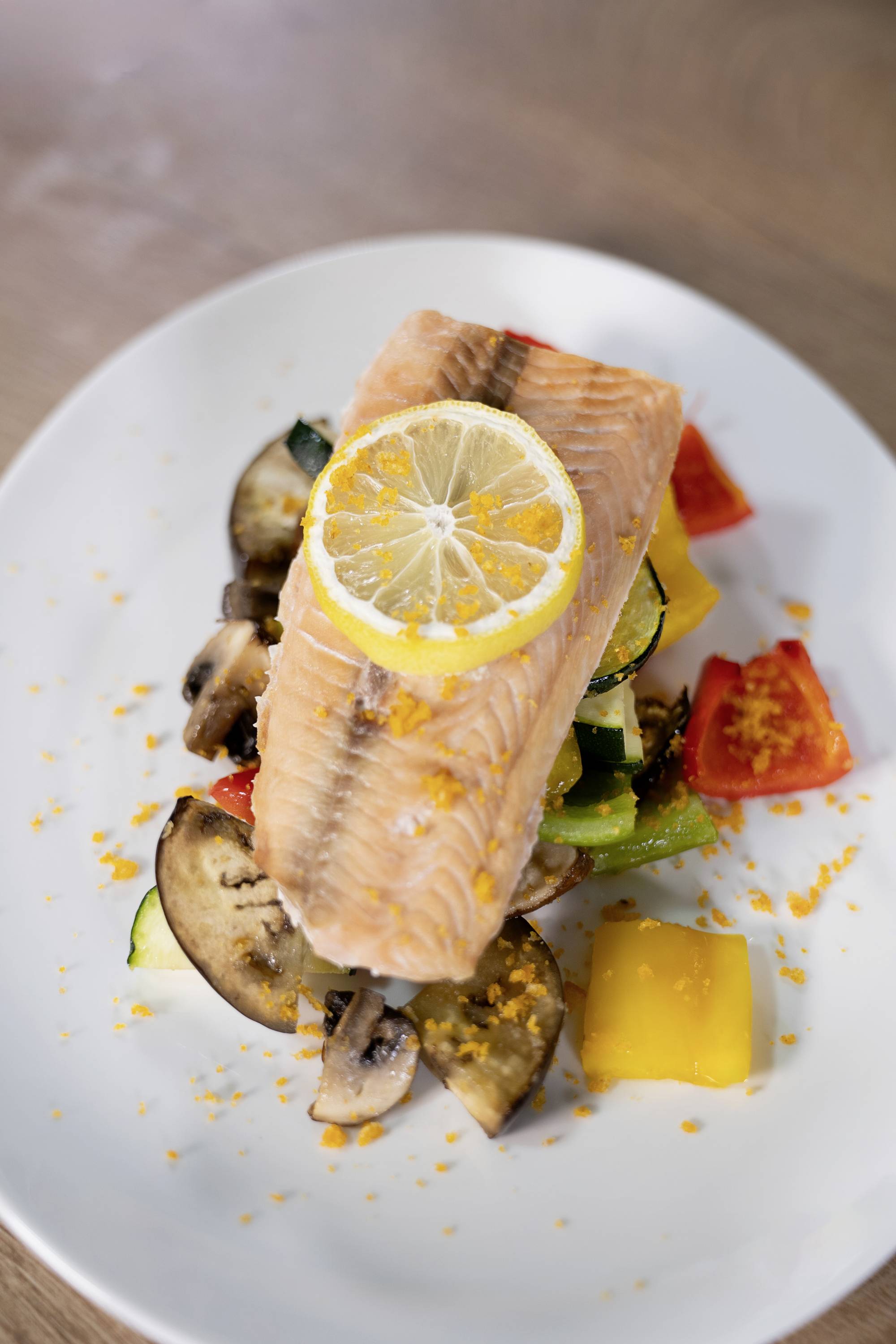 Filetto di salmone con fetta di limone su un letto di verdure come peperone, funghi e zucchine. Servito su un piatto bianco.