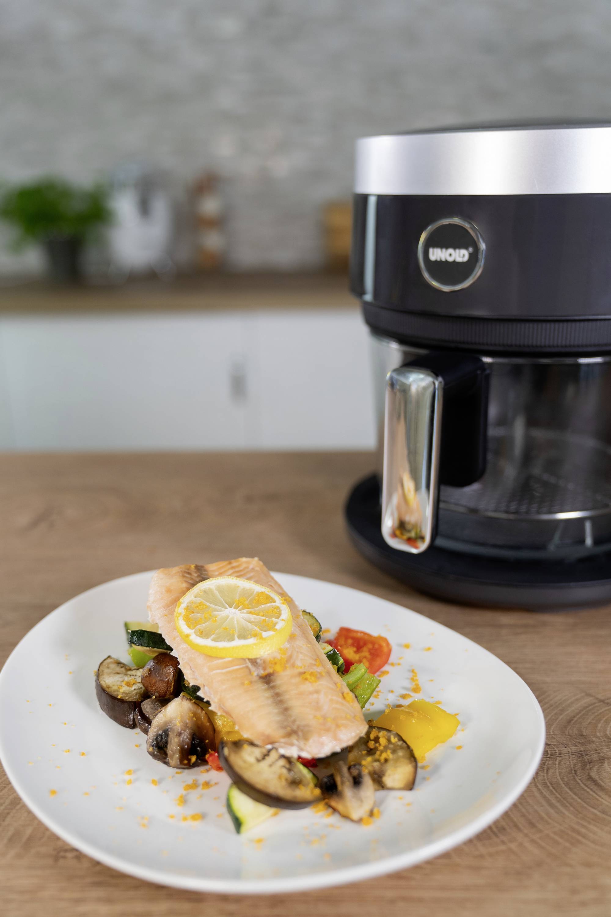 Un piatto con filetto di salmone alla griglia su verdure. Sullo sfondo, un robot da cucina nero è posizionato su un piano di lavoro.