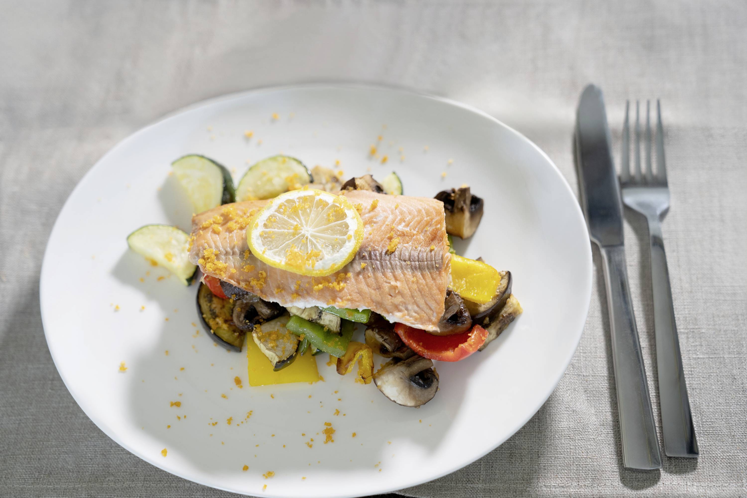 Un piatto di salmone fritto su un letto di zucchine, peperoni, pomodori e funghi, guarnito con una fetta di limone.