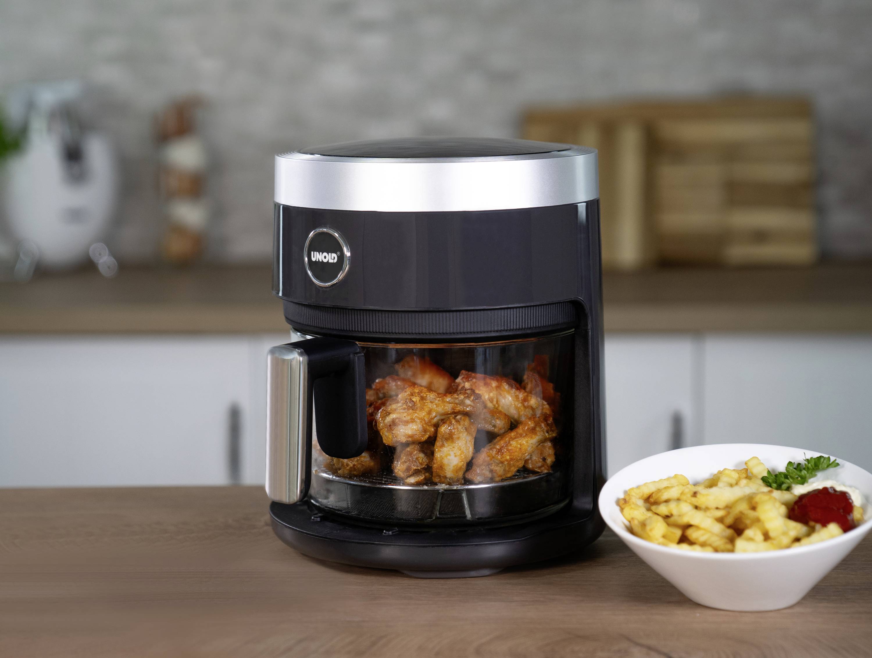Una friggitrice ad aria calda nera con cosce di pollo fritte su un piano da cucina. Accanto c'è una ciotola di patatine fritte.