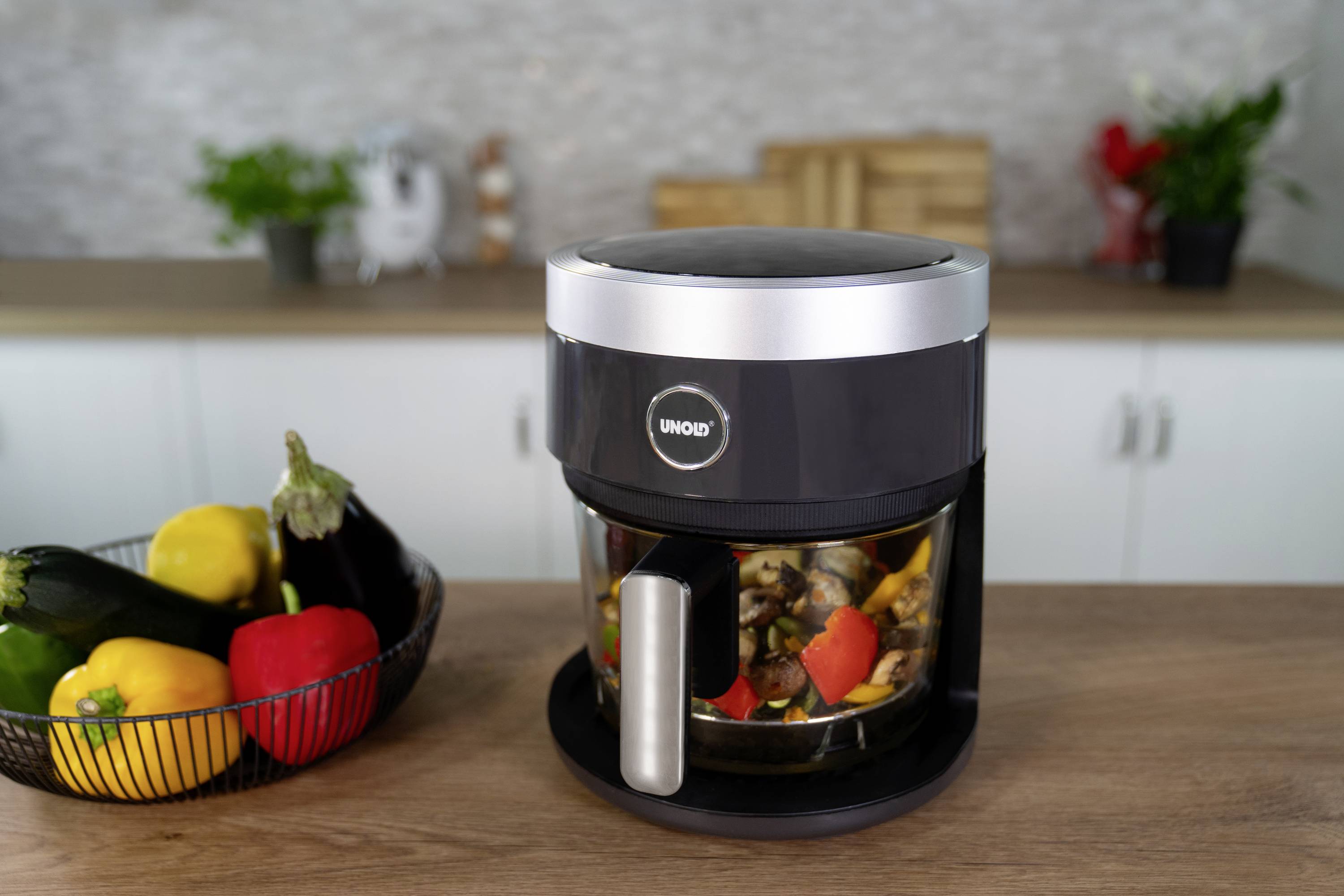 Un mixer nero è posizionato su un tavolo di legno accanto a un cestino pieno di verdure colorate come peperoni e melanzane.