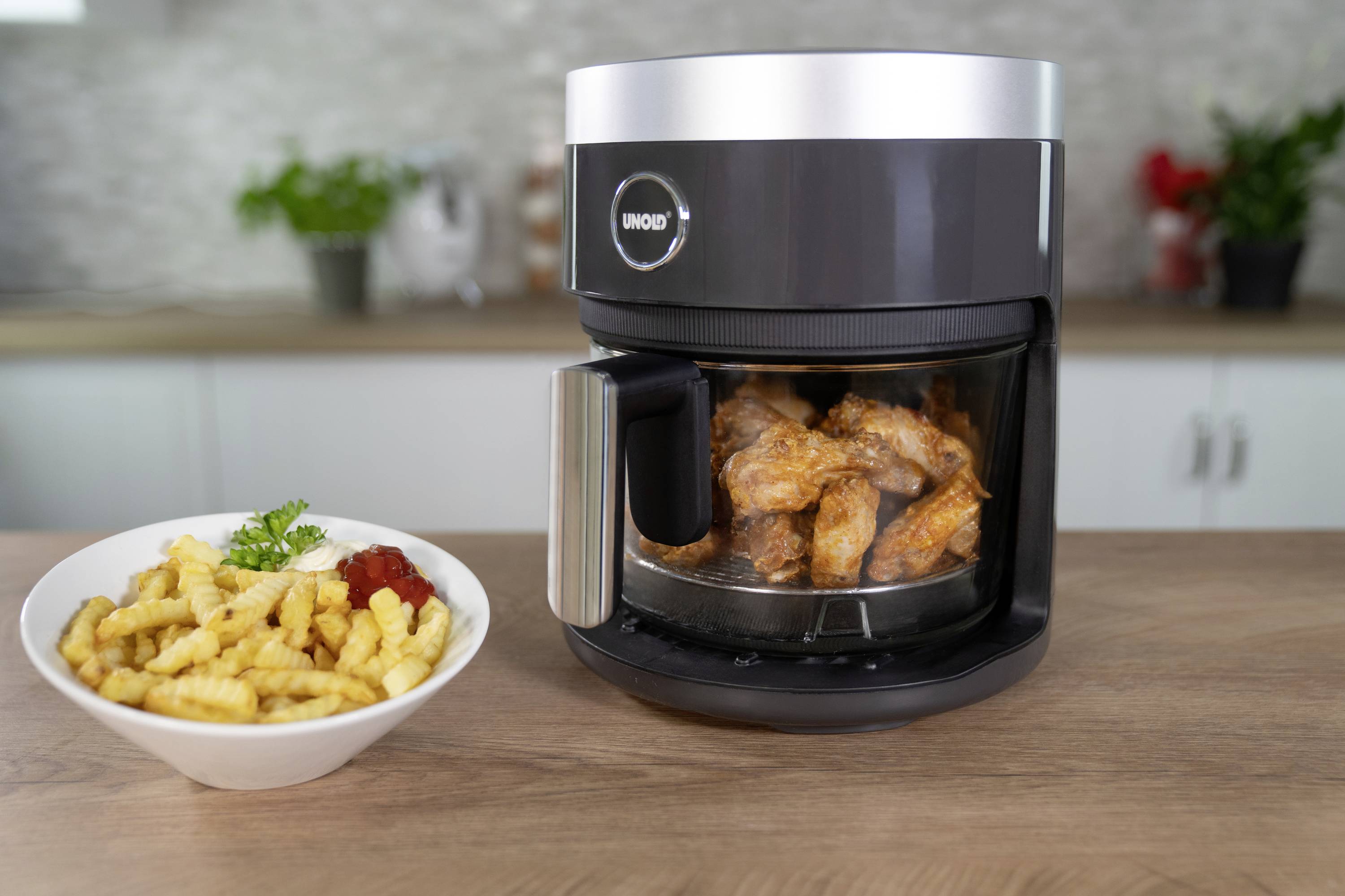 Una friggitrice ad aria calda riempita con ali di pollo è posizionata su un tavolo da cucina. Accanto, una ciotola di patatine fritte con ketchup.