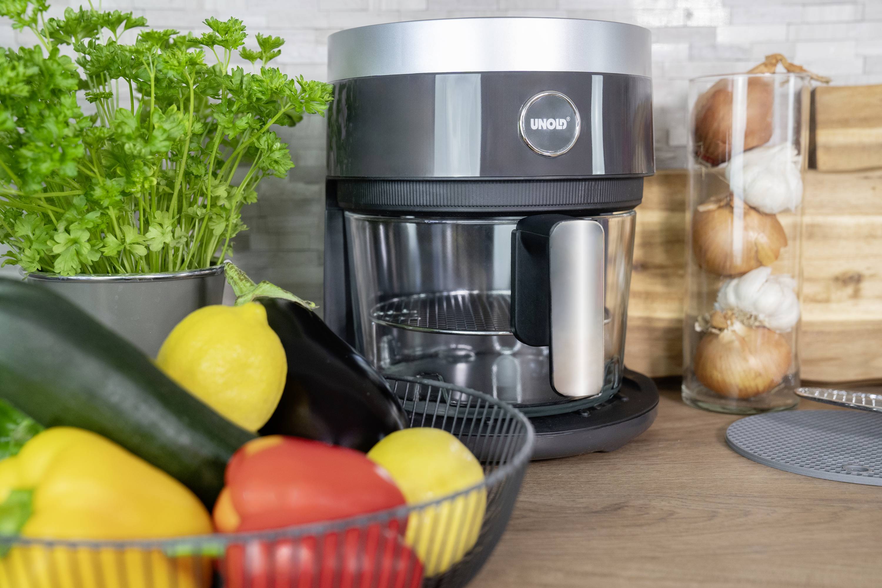 Robot da cucina sul piano di lavoro accanto a un cestino di verdure con peperoni e zucchine e una pentola con erbe fresche.