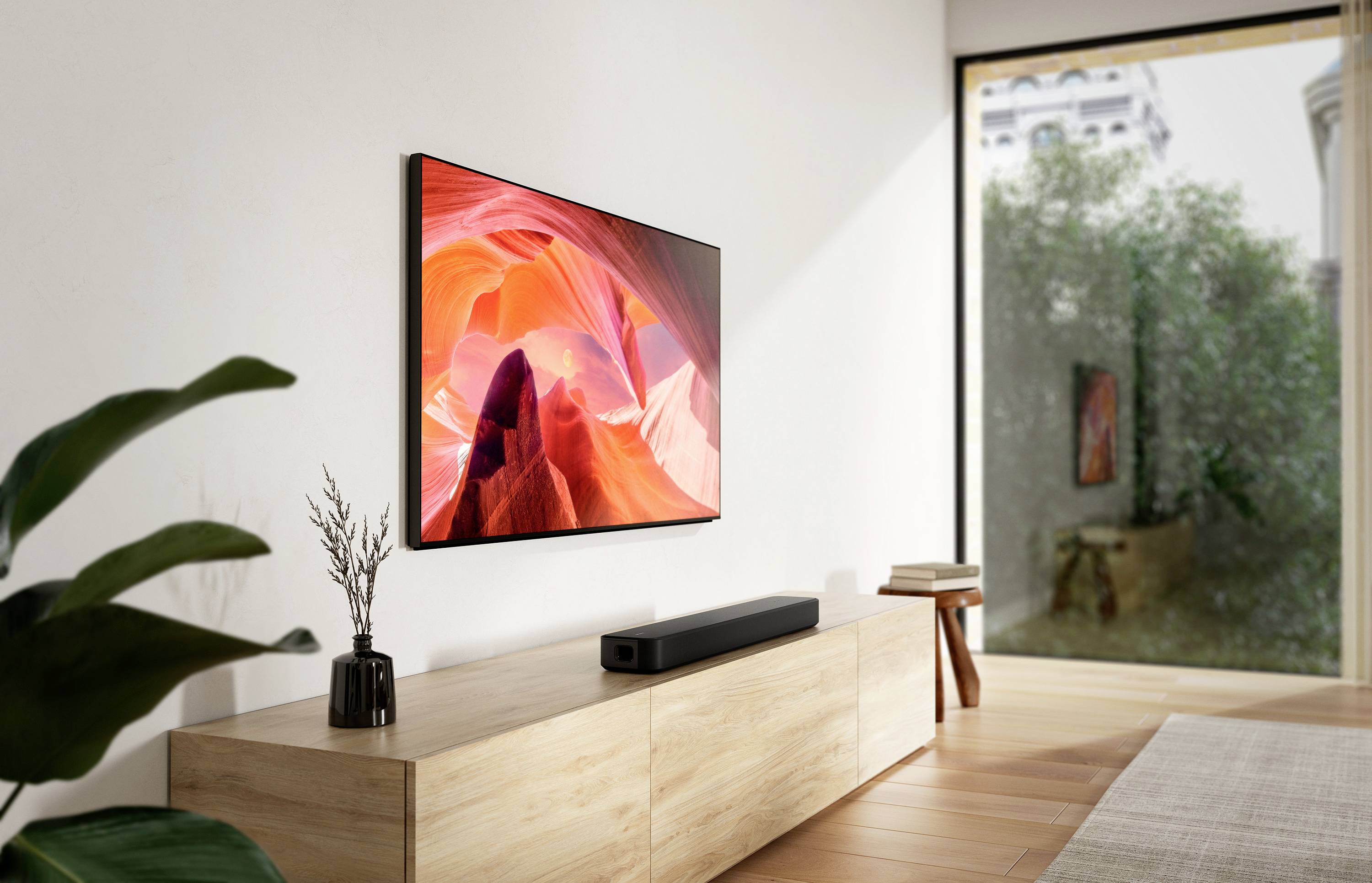Soggiorno con una console in legno, su cui è posizionata una soundbar. Sulla parete è appeso un grande schermo televisivo che mostra un motivo di montagna. Sullo sfondo, una grande finestra che si affaccia sul verde.