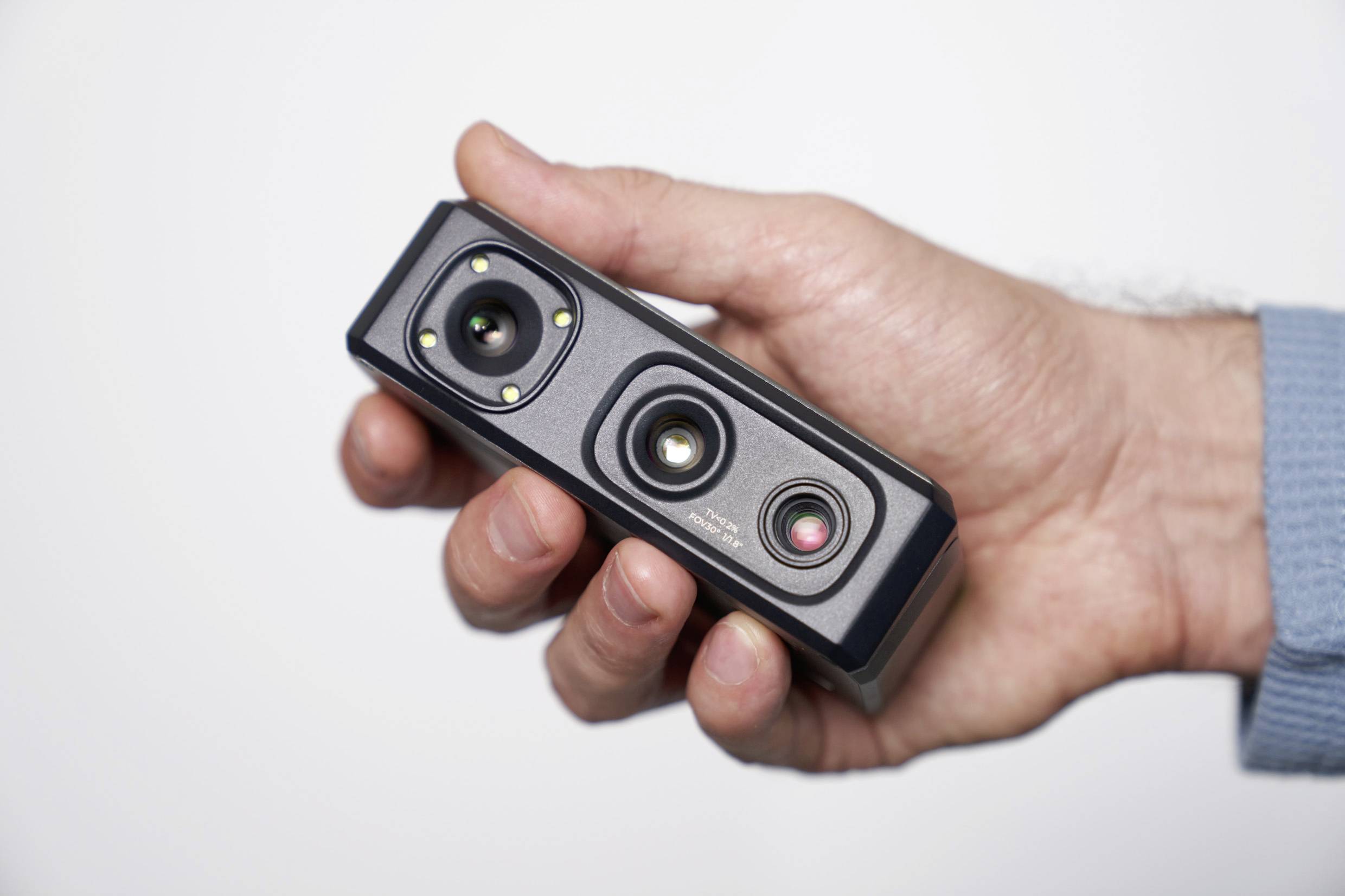Una mano tiene in mano una piccola fotocamera rettangolare con più lenti e sensori. La fotocamera è nera e ha una struttura compatta.