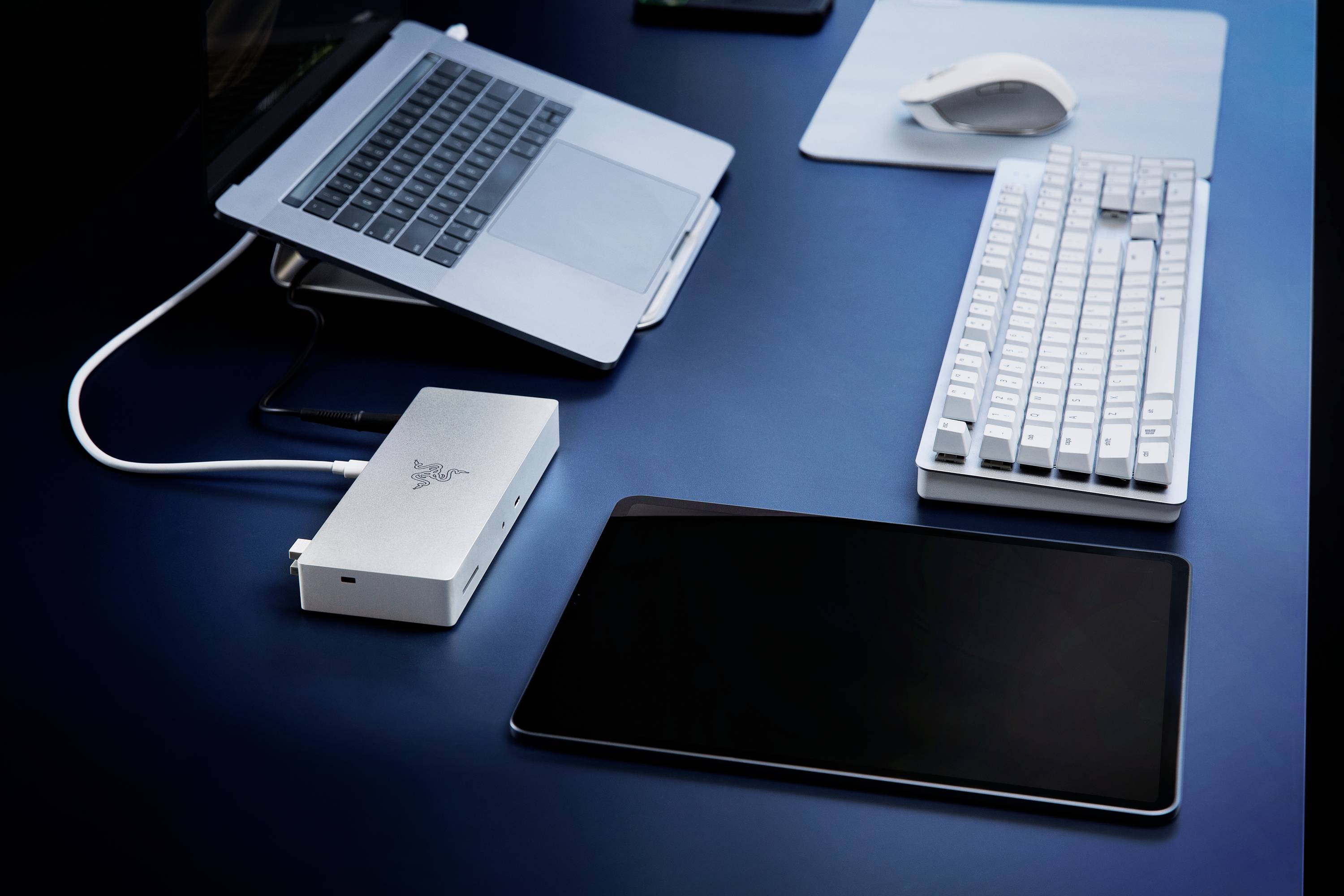 Un laptop, un hard disk esterno, una tastiera, un mouse e un tablet sono posizionati su un tavolo blu, collegati per lavorare.