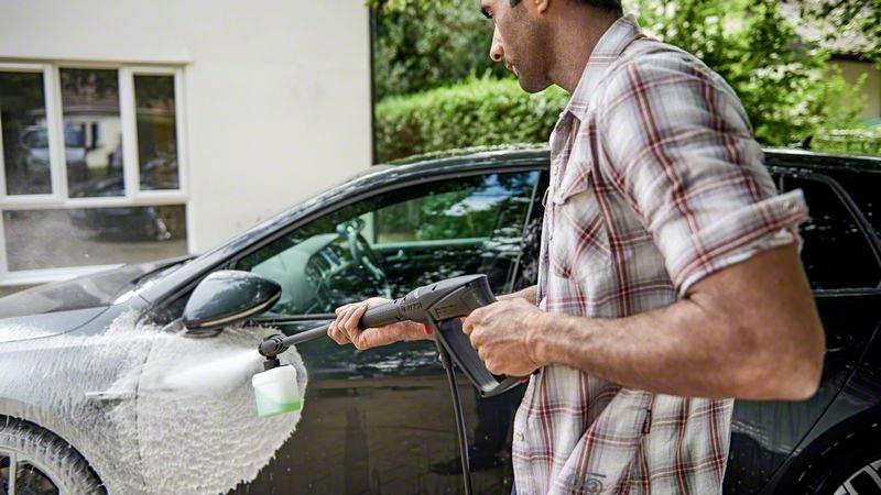 Una persona sta lavando un'auto nera con un'idropulitrice. L'auto è ricoperta di schiuma bianca. Sullo sfondo si vedono degli alberi.