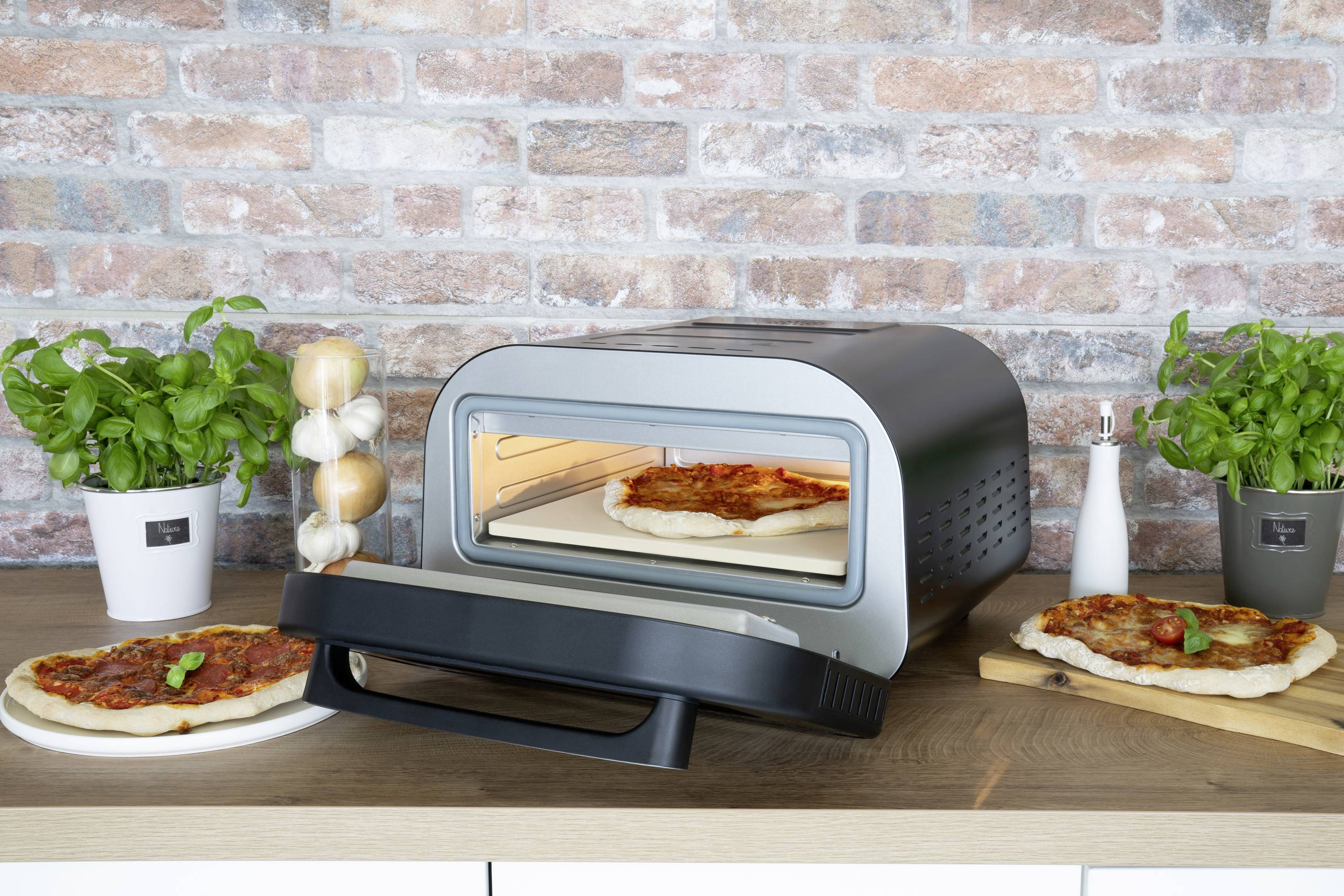 Un forno elettrico per pizza con la porta aperta, una pizza all'interno. Due pizze pronte accanto, circondante da erbe fresche e spezie.