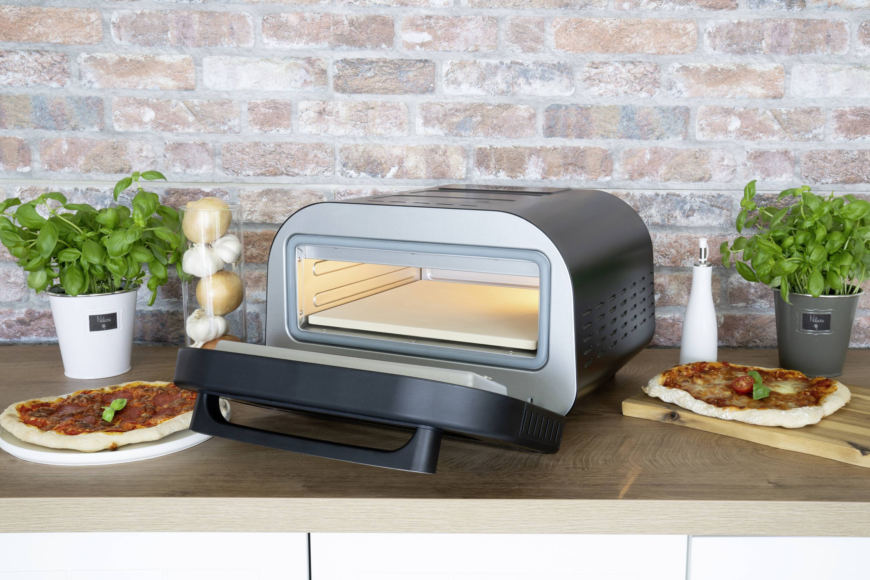 Un moderno forno per pizza su una linea di cucina, circondato da due pizze, erbe fresche e spezie, davanti a un muro di mattoni.