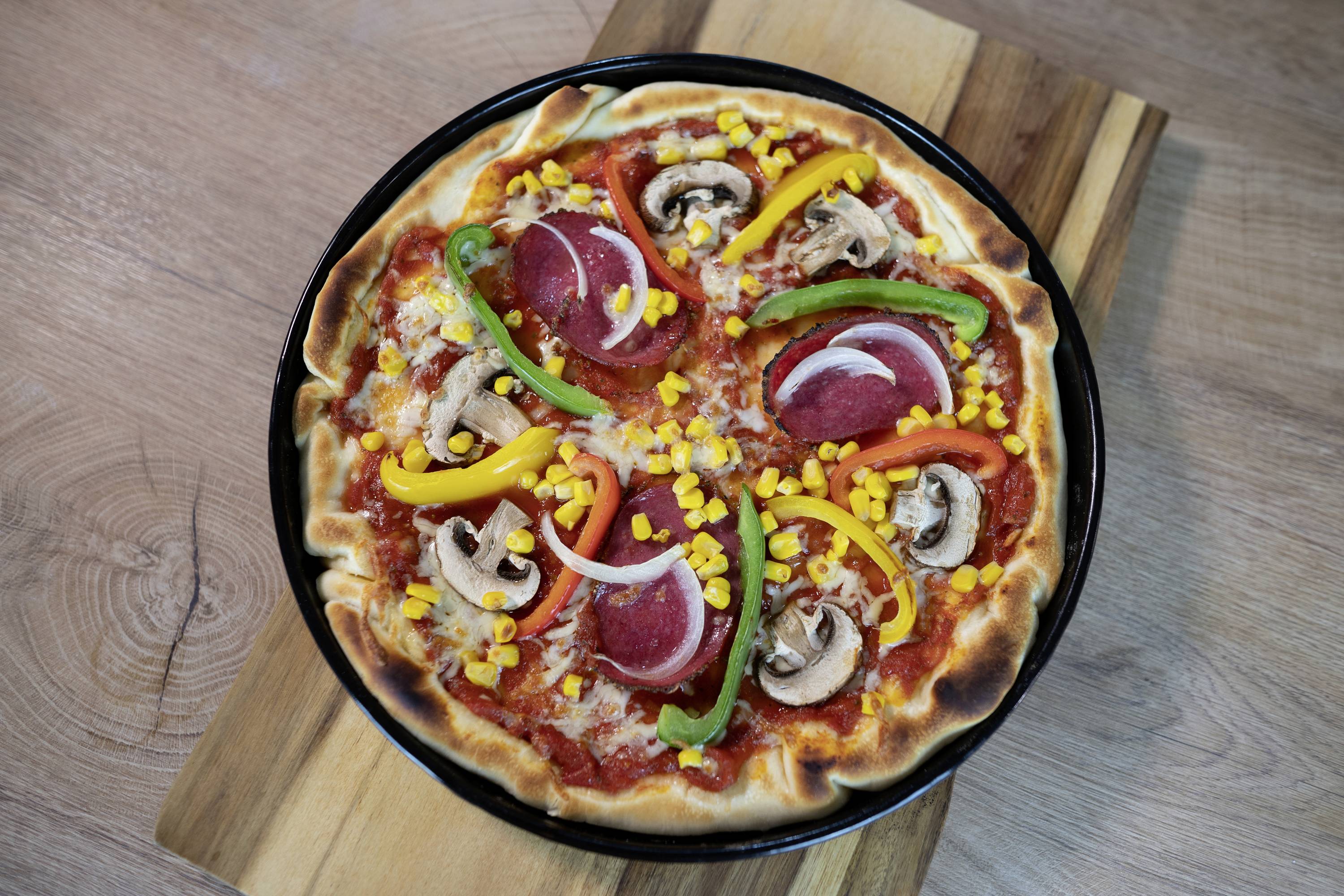 Pizza con salsa di pomodoro, peperoni, cipolle rosse, peperone, mais e funghi su un tagliere di legno.