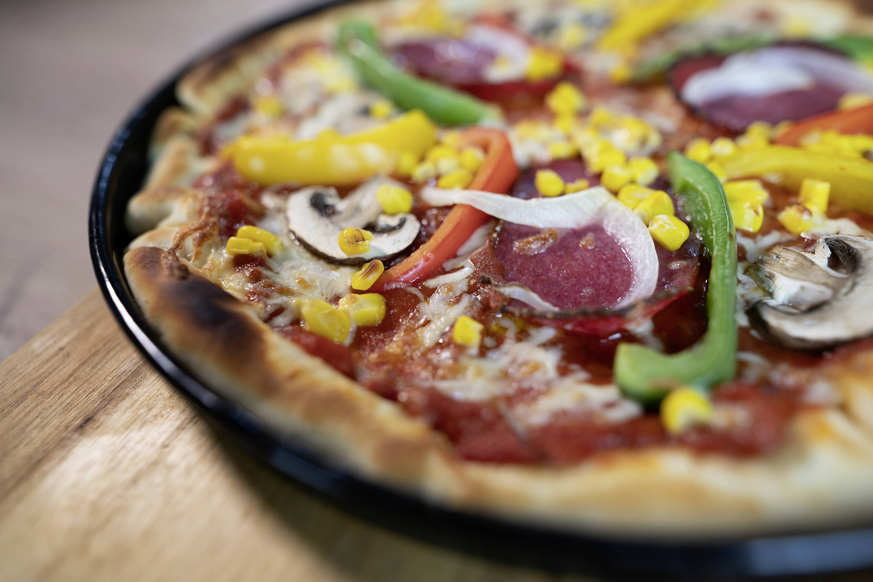 Pizza con salame, peperone, mais e funghi su un tagliere di legno.