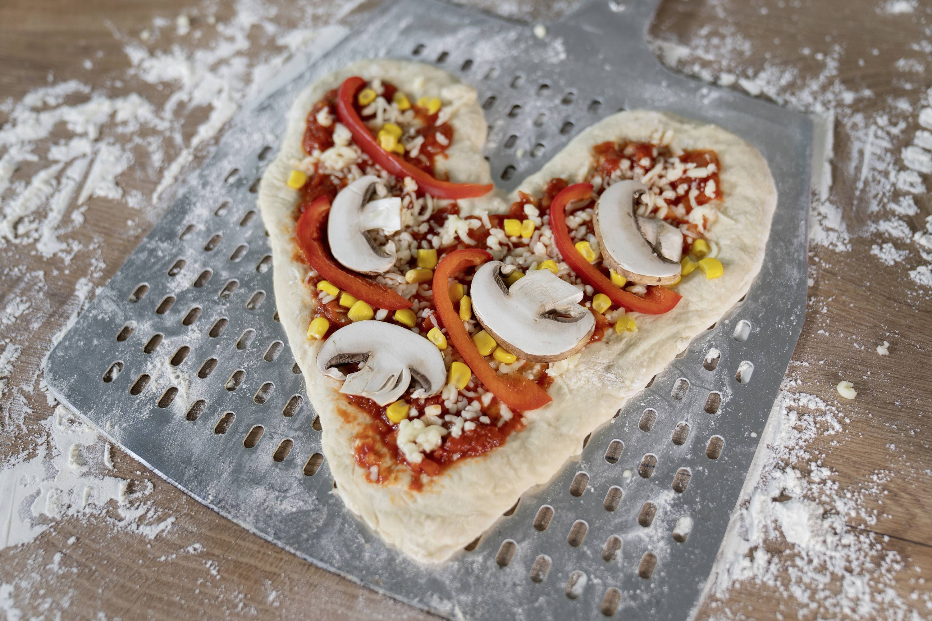 Pizza a forma di cuore con salsa di pomodoro, strisce di peperone, funghi, mais e noci su una pala per pizza.