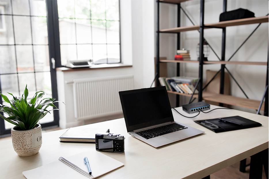 Una scrivania con un laptop, una macchina fotografica, un quaderno e una pianta in un ufficio moderno con scaffali e grandi finestre.
