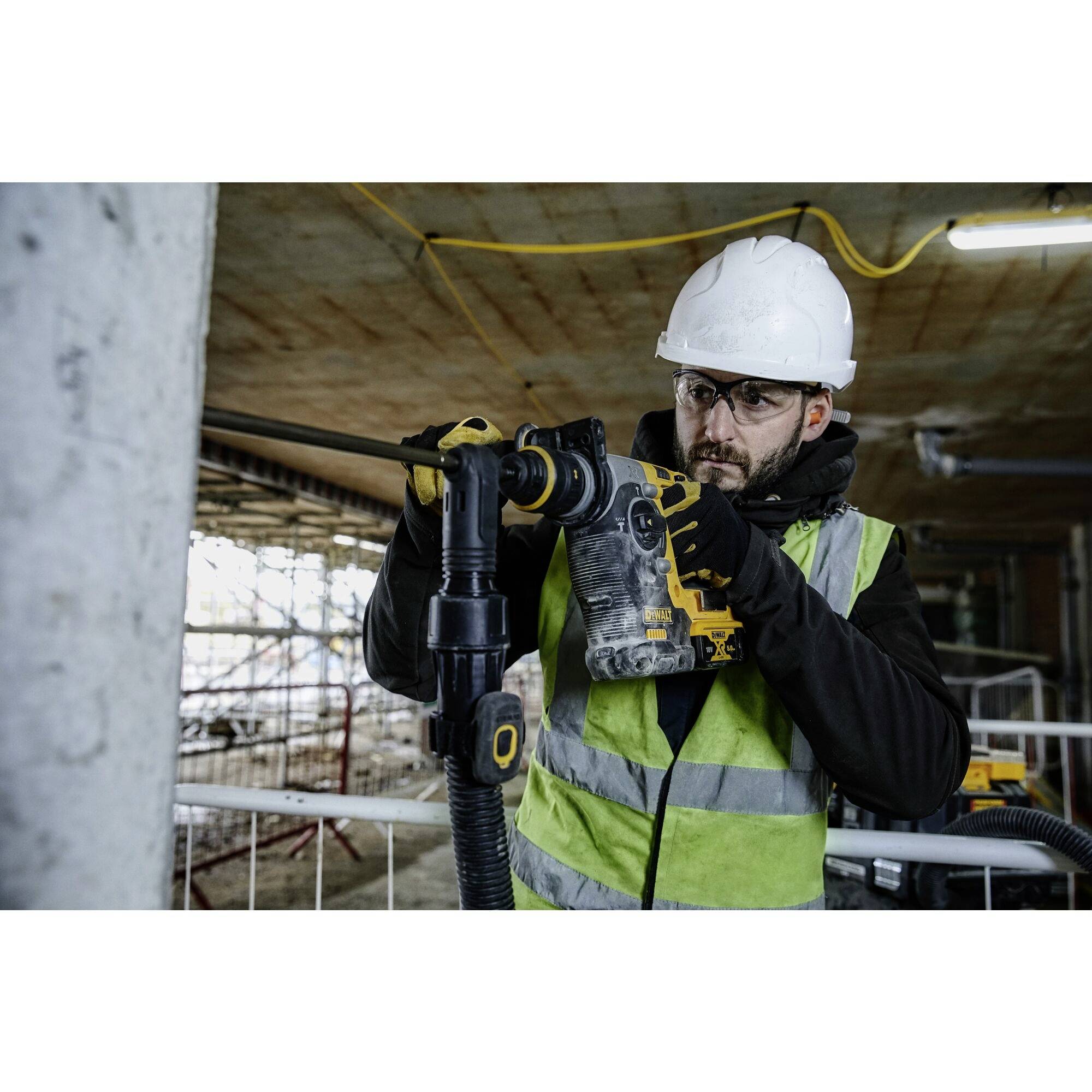 Un operaio edile indossa indumenti protettivi e utilizza un utensile elettrico in un cantiere. Sullo sfondo sono visibili dei ponteggi.